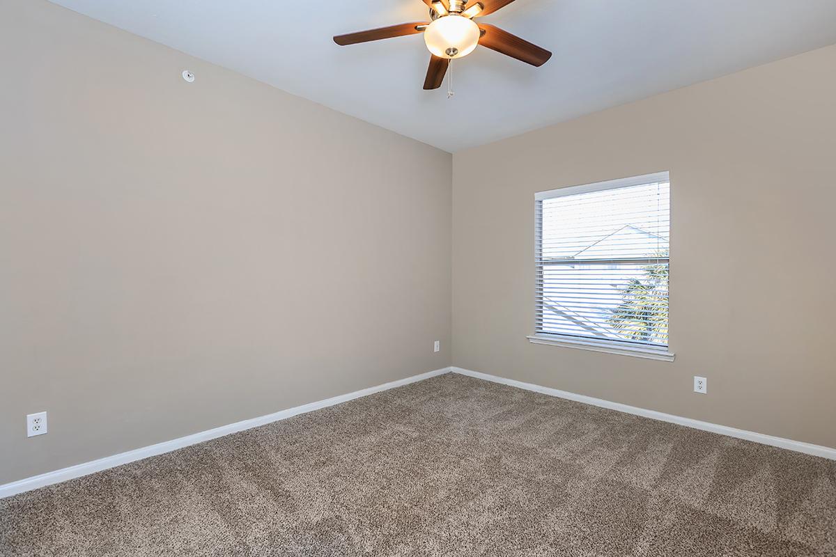 A vacant room with light beige walls and a ceiling fan. The floor is covered with light brown carpet. A window with horizontal blinds lets in natural light, revealing a view of outside greenery. The room is empty, providing a neutral space for various furnishings.