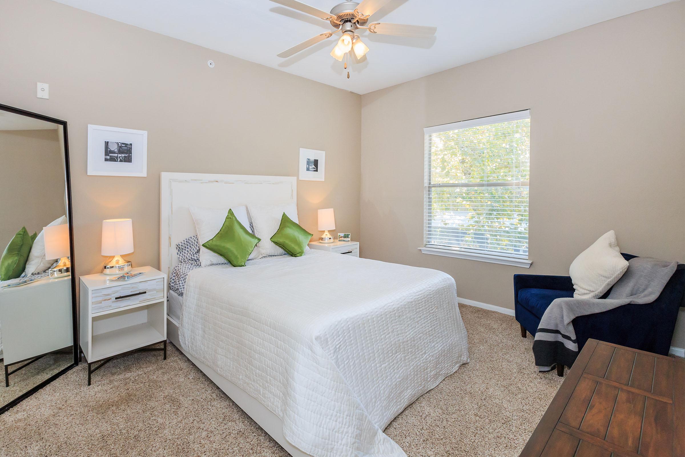 Cozy bedroom with a queen-sized bed topped with white bedding and green accent pillows. A large mirror is positioned next to the bed, complemented by two bedside lamps. A window allows natural light in, while a small blue armchair sits in the corner, and a wooden table adds warmth to the decor.