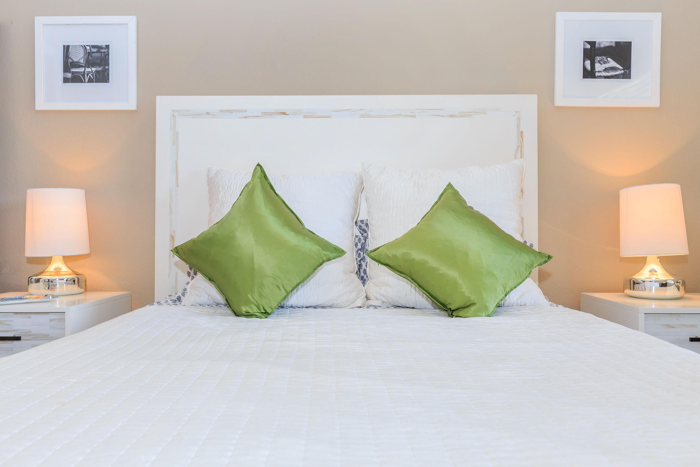 A cozy bedroom setting featuring a white bed with two plush green square pillows. On either side of the bed are two bedside tables with lamps. The wall above has two framed black-and-white photographs, adding a touch of decor to the warm, inviting space.