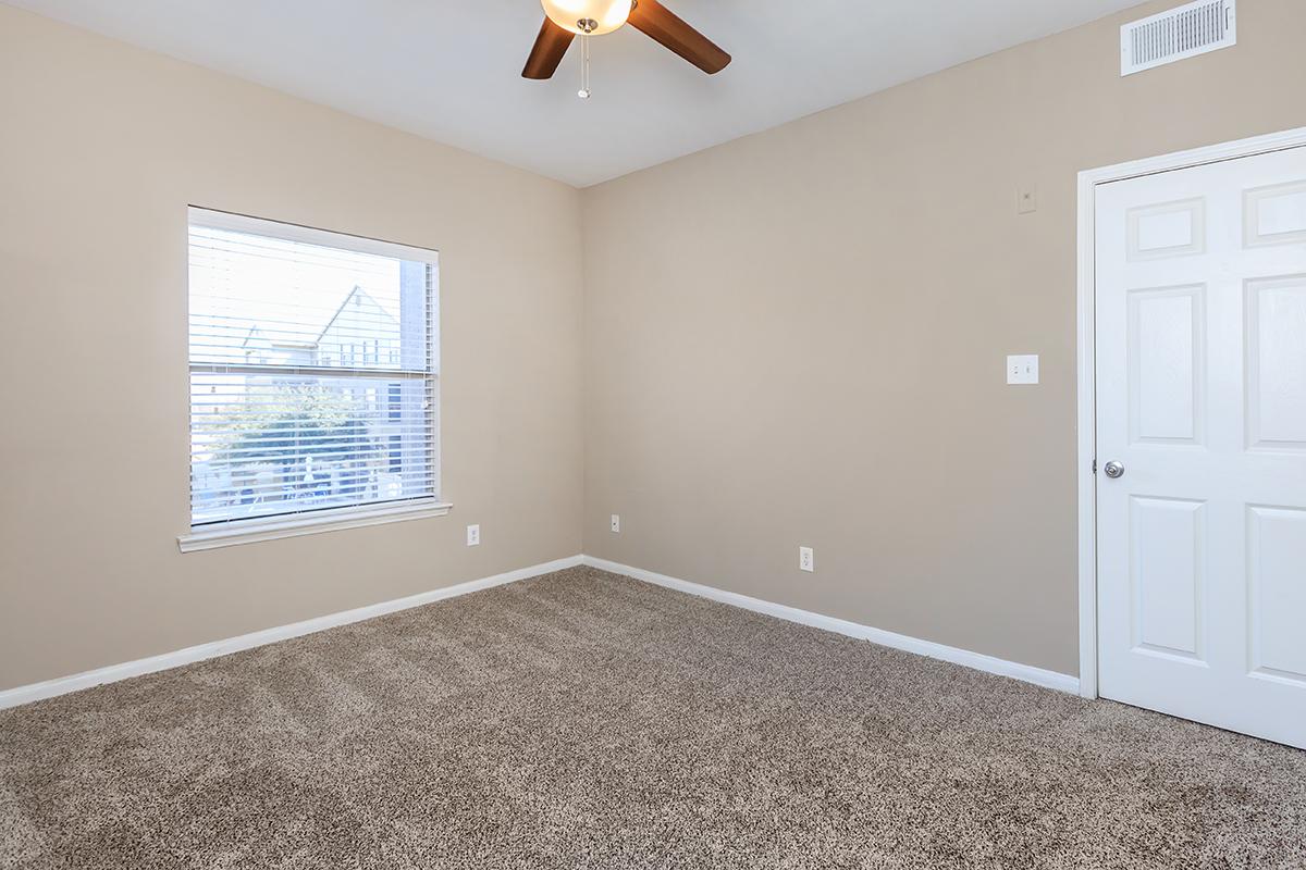 A sparsely furnished room with light beige walls and plush carpet. A ceiling fan is present, and there's a single window with blinds allowing natural light. A white door is visible on the right, completing the simple, neutral aesthetic of the space.