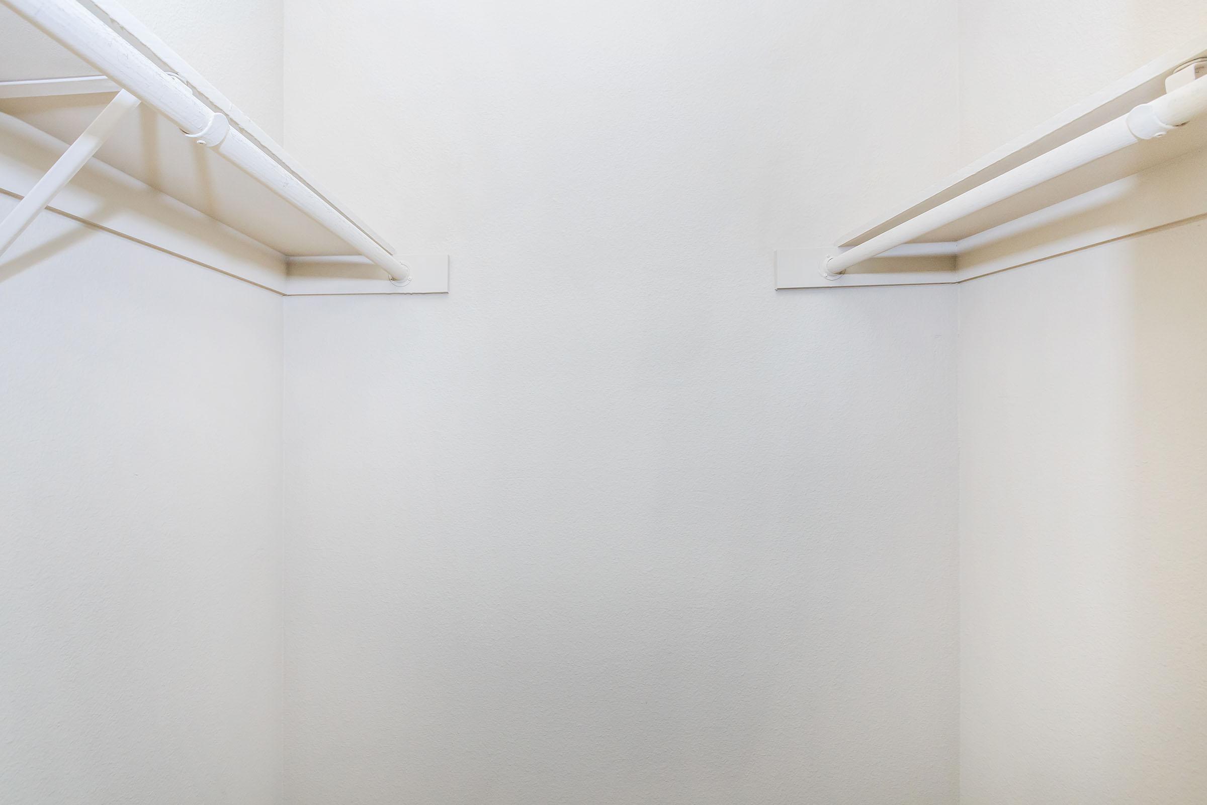 An empty closet with a plain white wall and two white hanging rods. The space has minimal decoration, creating a clean and simple appearance. The lighting is neutral, emphasizing the uncluttered nature of the area.