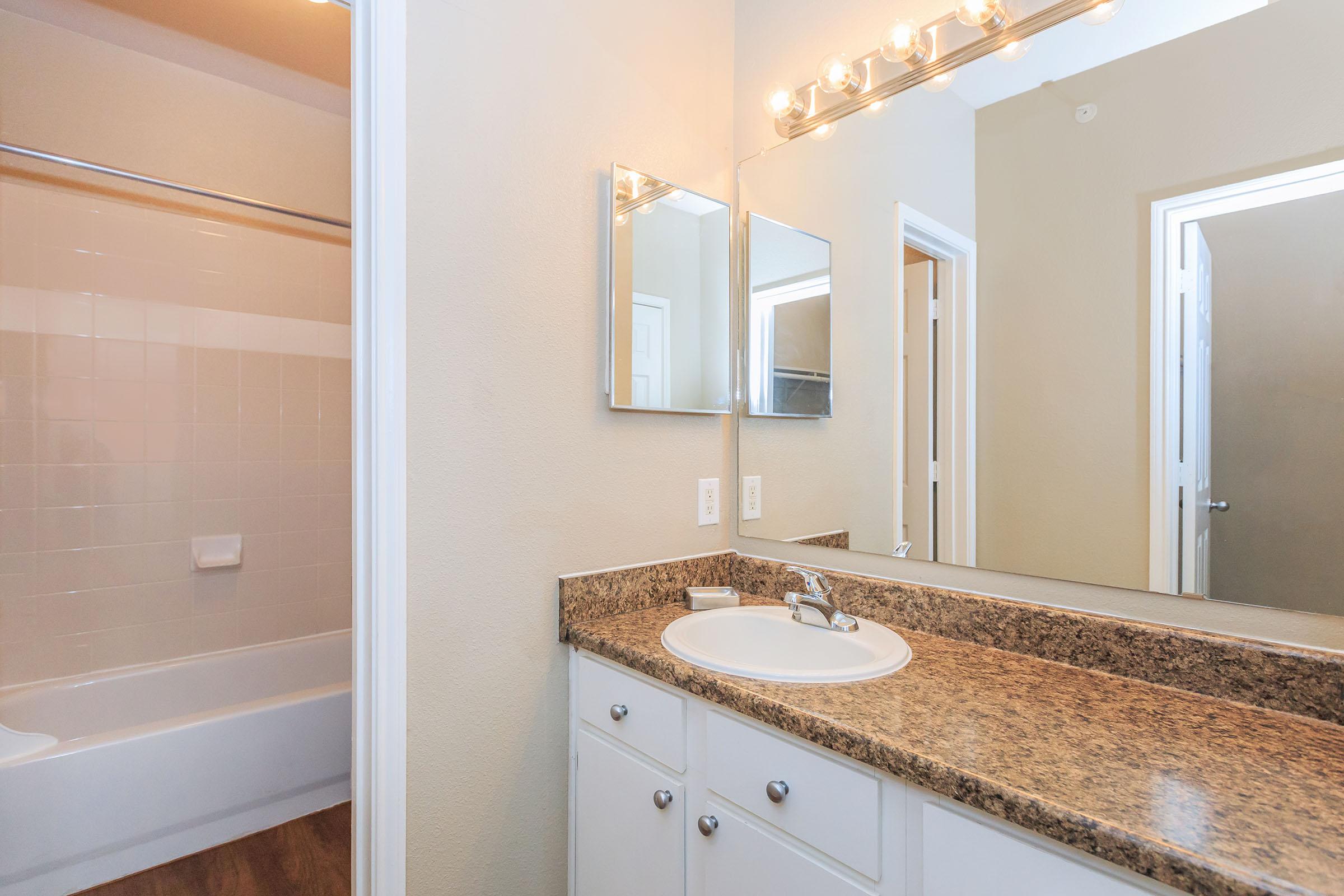 A well-lit bathroom featuring a granite countertop with a sink and a large mirror. The space includes a white bathtub and shower combo, light-colored walls, and a door leading to another room, creating a clean and modern aesthetic.