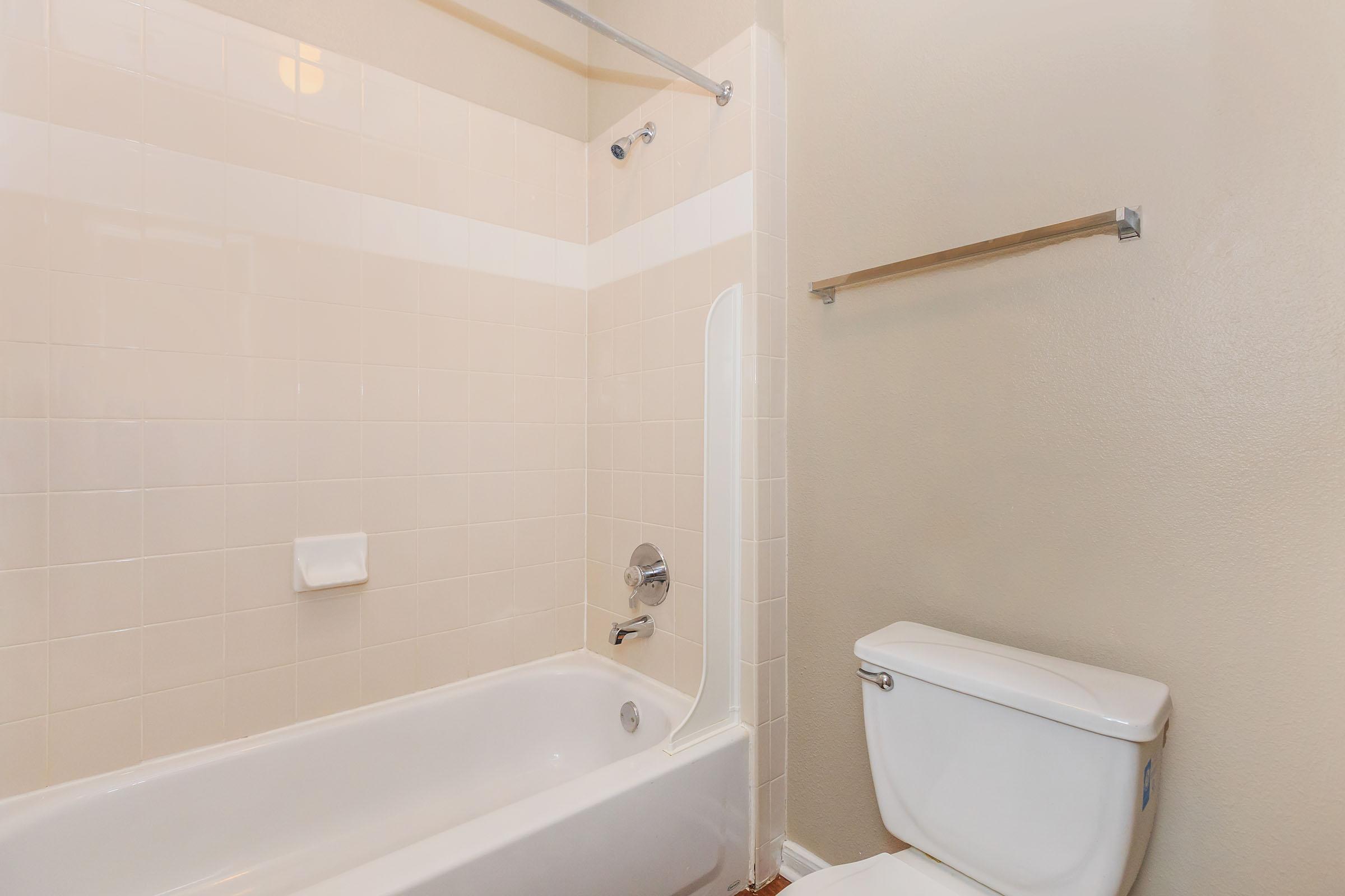 A clean, simple bathroom featuring a white bathtub with a showerhead, beige tiled walls, and a toilet. A towel bar is mounted on the wall beside the bathtub. The overall color scheme is light and neutral, creating a spacious feel.