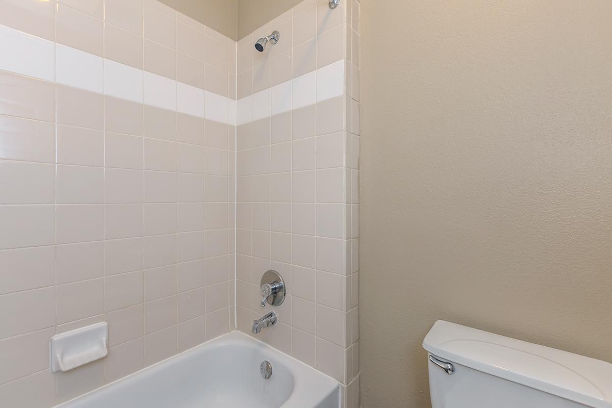 A clean bathroom featuring a bathtub with an attached shower head, white tiled walls, and a toilet nearby. The tiles are light beige with a horizontal white stripe. The space is simple and well-maintained, reflecting a modern, minimalistic design.