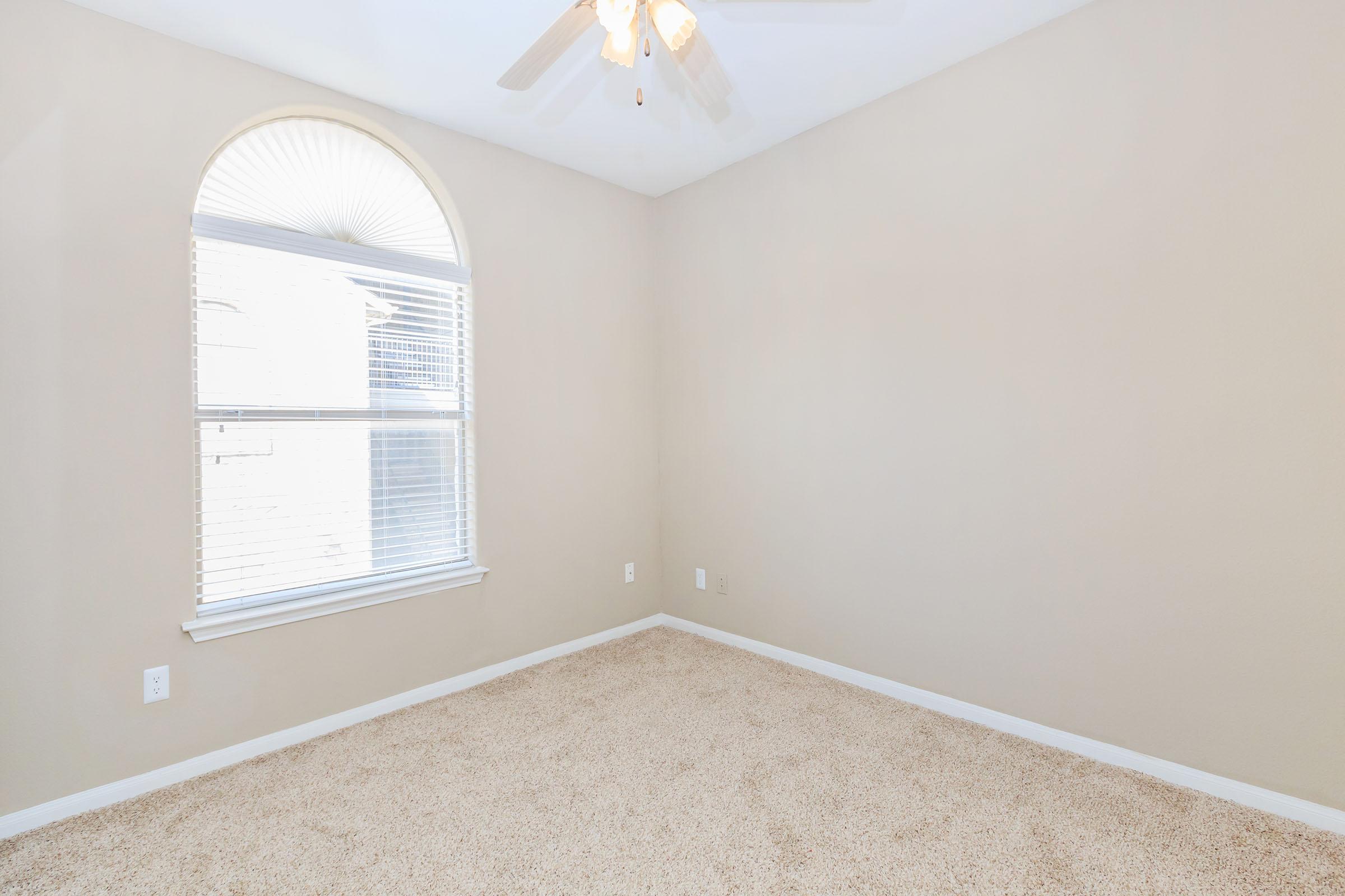 Empty room with beige walls and carpet, featuring a large window with blinds and a decorative arch at the top. A ceiling fan is visible, and there are no furniture or decorations in the space, creating a clean and minimalist appearance.