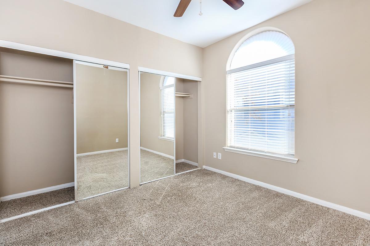 A well-lit bedroom featuring beige walls and carpet, with a large window on one side. There are mirrored sliding closet doors reflecting the room, enhancing the sense of space. The ceiling has a ceiling fan, adding to the comfort of the environment.