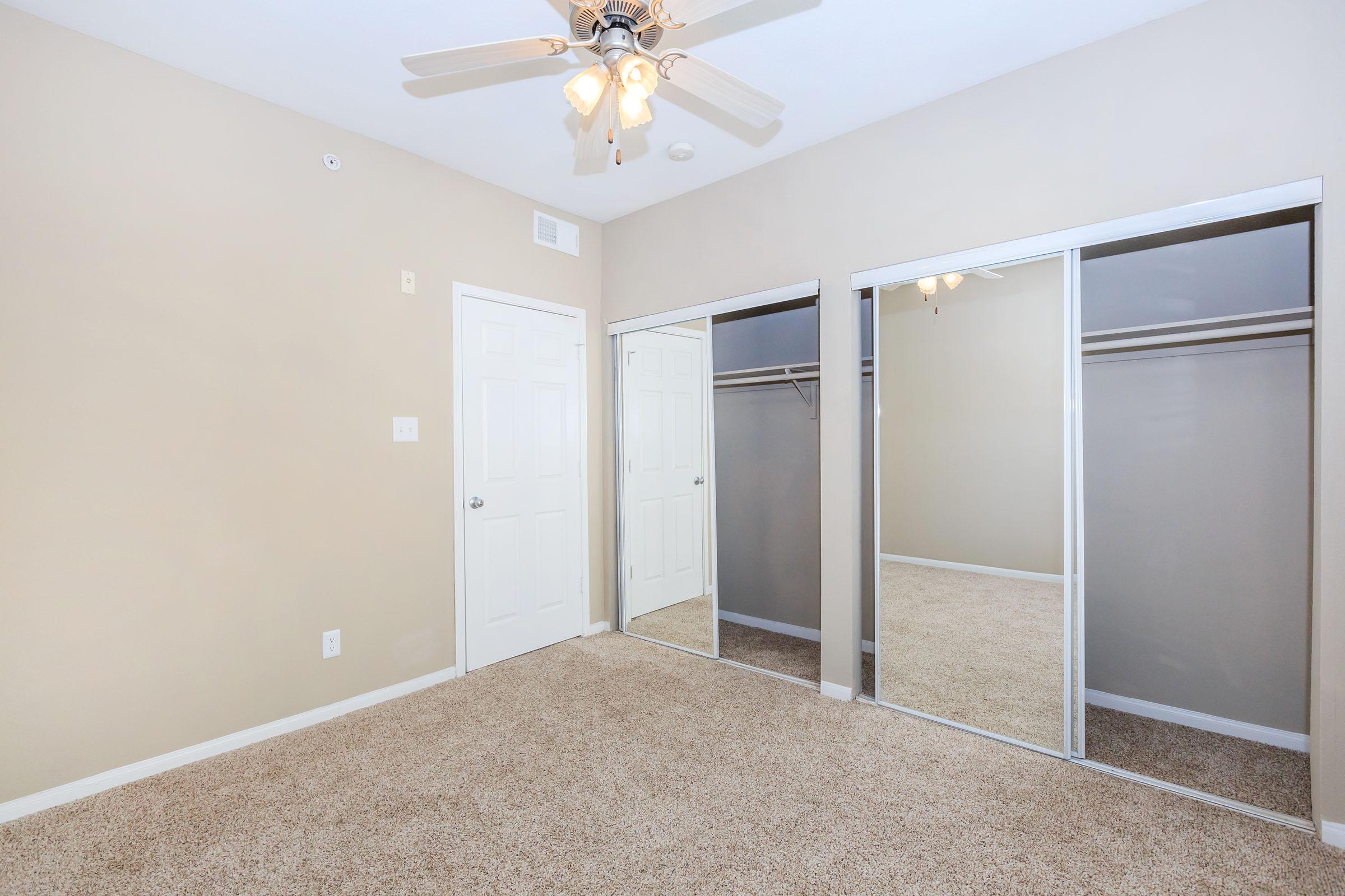 A spacious, empty bedroom featuring light beige walls and carpet. The room has a ceiling fan with light fixtures and a mirrored sliding door closet. There’s a white door leading to another area, and the overall space is well-lit and inviting.