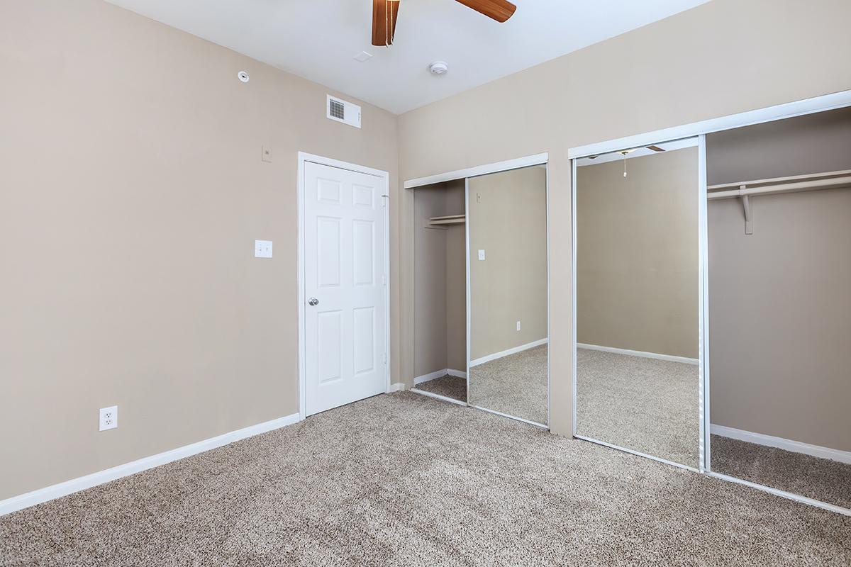 A neutral-colored bedroom featuring light brown walls, a ceiling fan, and carpeted flooring. Two mirrored closet doors are visible, reflecting a plain space. A white door leads to another area, and there are no furnishings in the room.