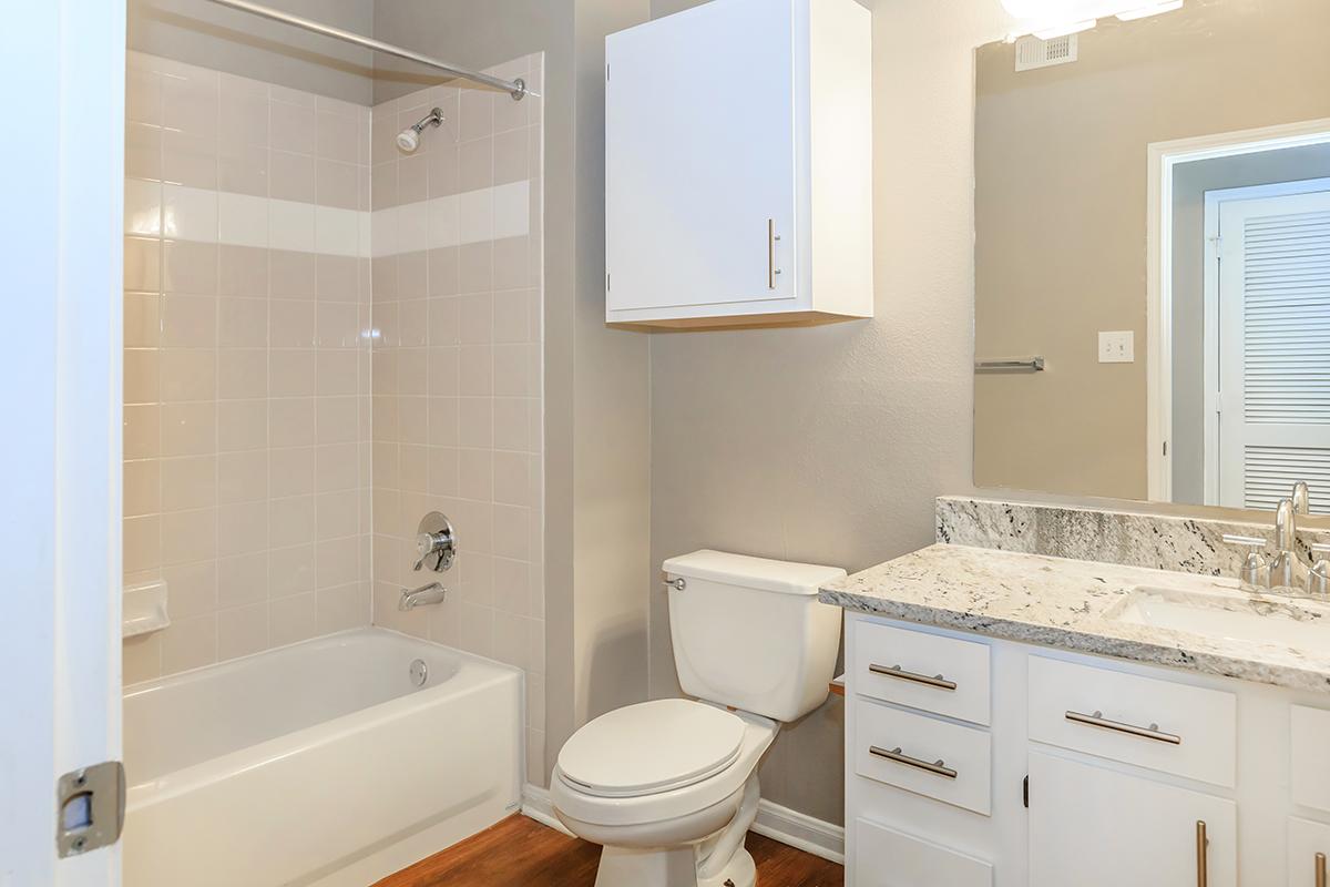 A clean bathroom featuring a shower/tub combo with white tiles, a modern vanity with a granite countertop, and a white toilet. The walls are painted light gray, and there's a cabinet above the sink for storage. Hardwood flooring adds warmth to the space.