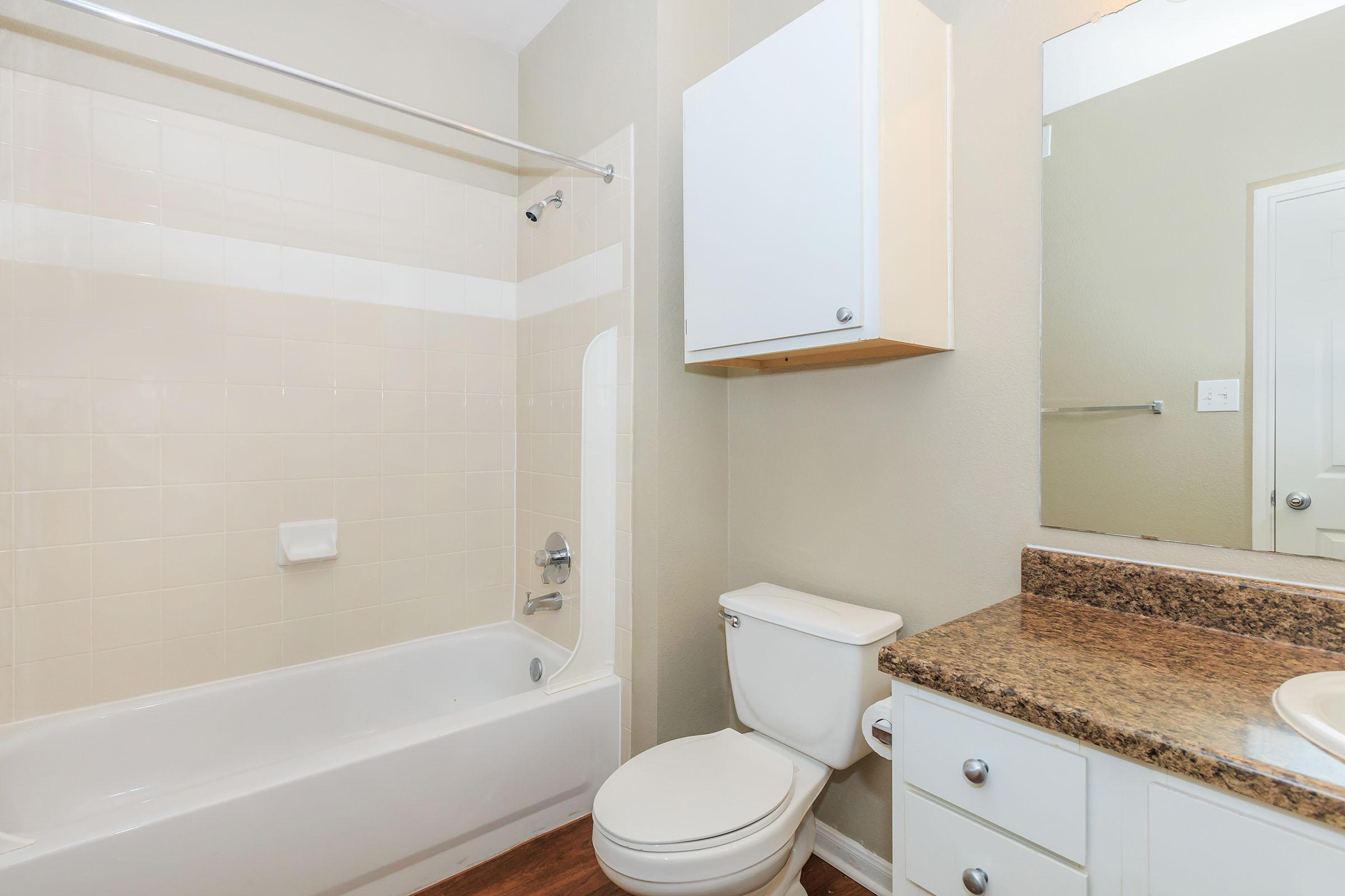 A clean, well-lit bathroom featuring a bathtub with a shower, a white toilet, and a granite countertop with a sink. The walls are painted a light color, and there is a cabinet mounted above the toilet for storage. The floor is a warm, wood-like material, adding a cozy touch to the space.