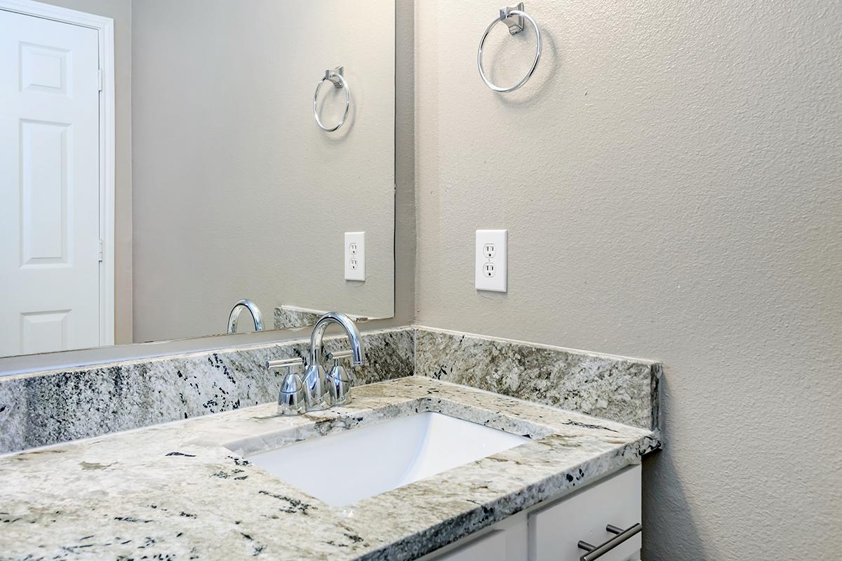 A modern bathroom countertop features a white sink and sleek chrome faucet, with a textured granite surface. The wall is a soft gray, and there's a round towel ring mounted nearby. In the background, a white door is slightly ajar, adding a touch of depth to the space. A mirror reflects the clean lines of the design.