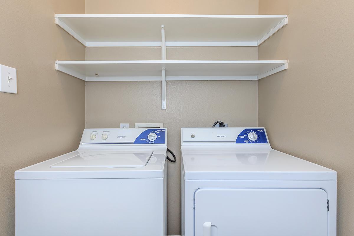 A clean laundry room featuring a white washing machine and a white dryer side by side. There are two white shelves above the appliances, with a plain beige wall in the background. The image conveys a tidy and organized space for laundry tasks.