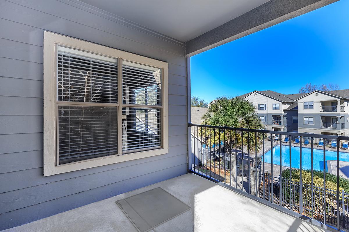 View from a balcony overlooking a pool area. The balcony features a gray wall with a window that has white blinds. In the background, there are palm trees and several apartment buildings, all under a clear blue sky. The scene conveys a relaxed, sunny atmosphere.