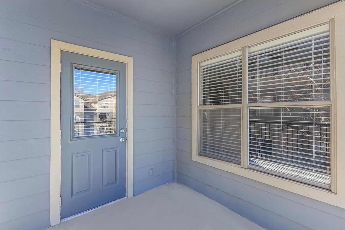 A gray exterior entryway featuring a blue door on the left and a large window on the right with white blinds. The walls are painted light gray, and the space is well-lit with natural light coming through the window. The floor is a light-colored concrete.