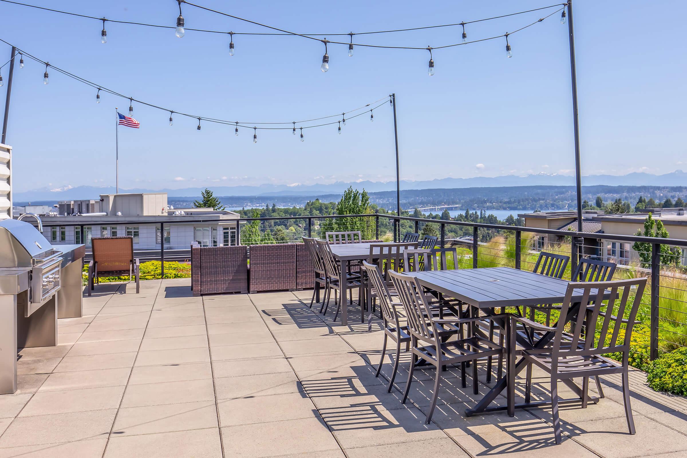 A rooftop patio featuring multiple dining tables and chairs, string lights overhead, with a view of a lake and mountains in the background. An American flag is visible in the distance, and there is a grill area to the side. The setting is sunny and well-maintained, creating an inviting outdoor space.