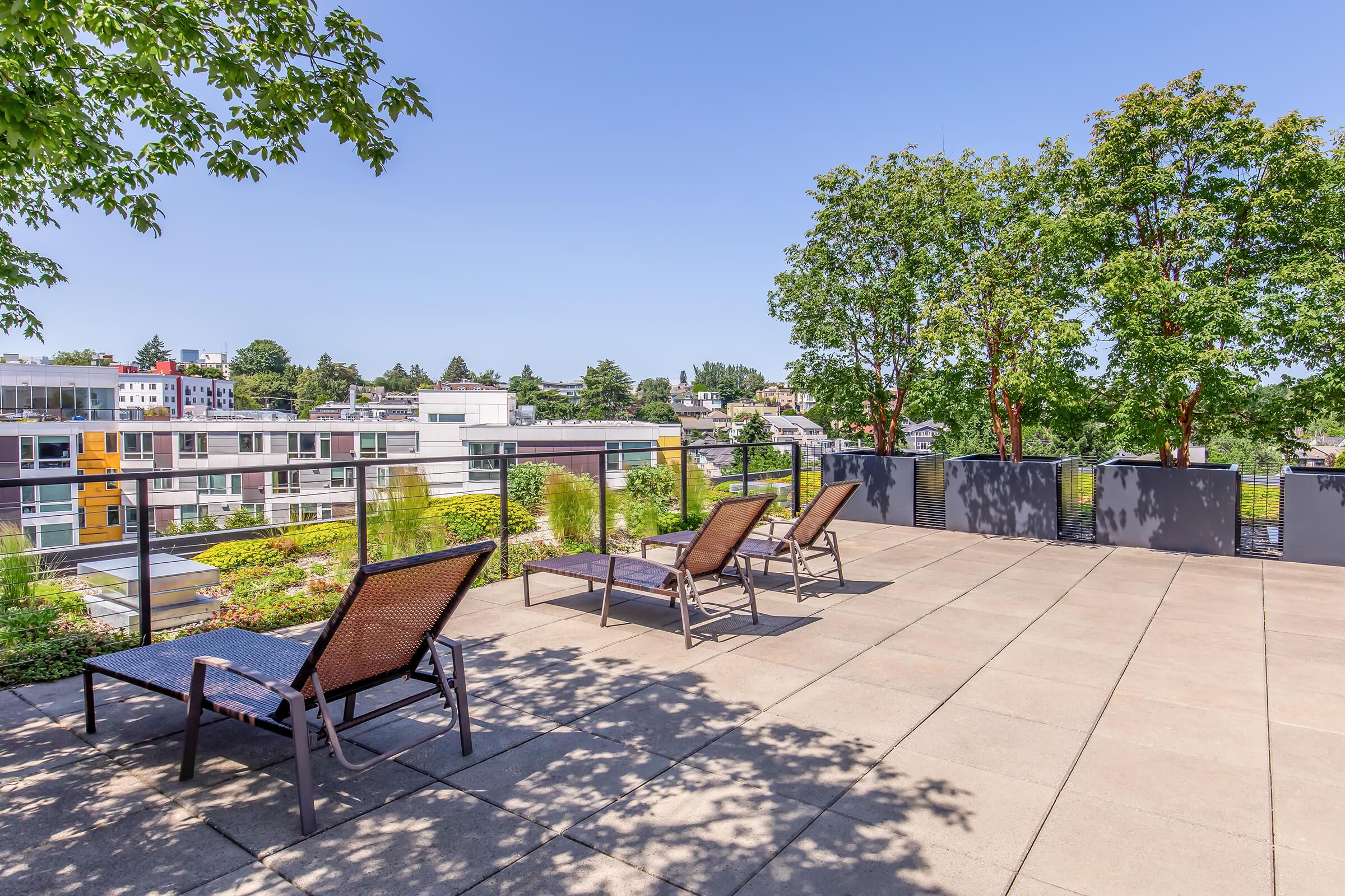 A rooftop terrace featuring several lounge chairs, surrounded by greenery and trees. The setting offers a clear blue sky and a view of modern buildings in the background. The area is designed for relaxation and enjoyment of outdoor spaces.