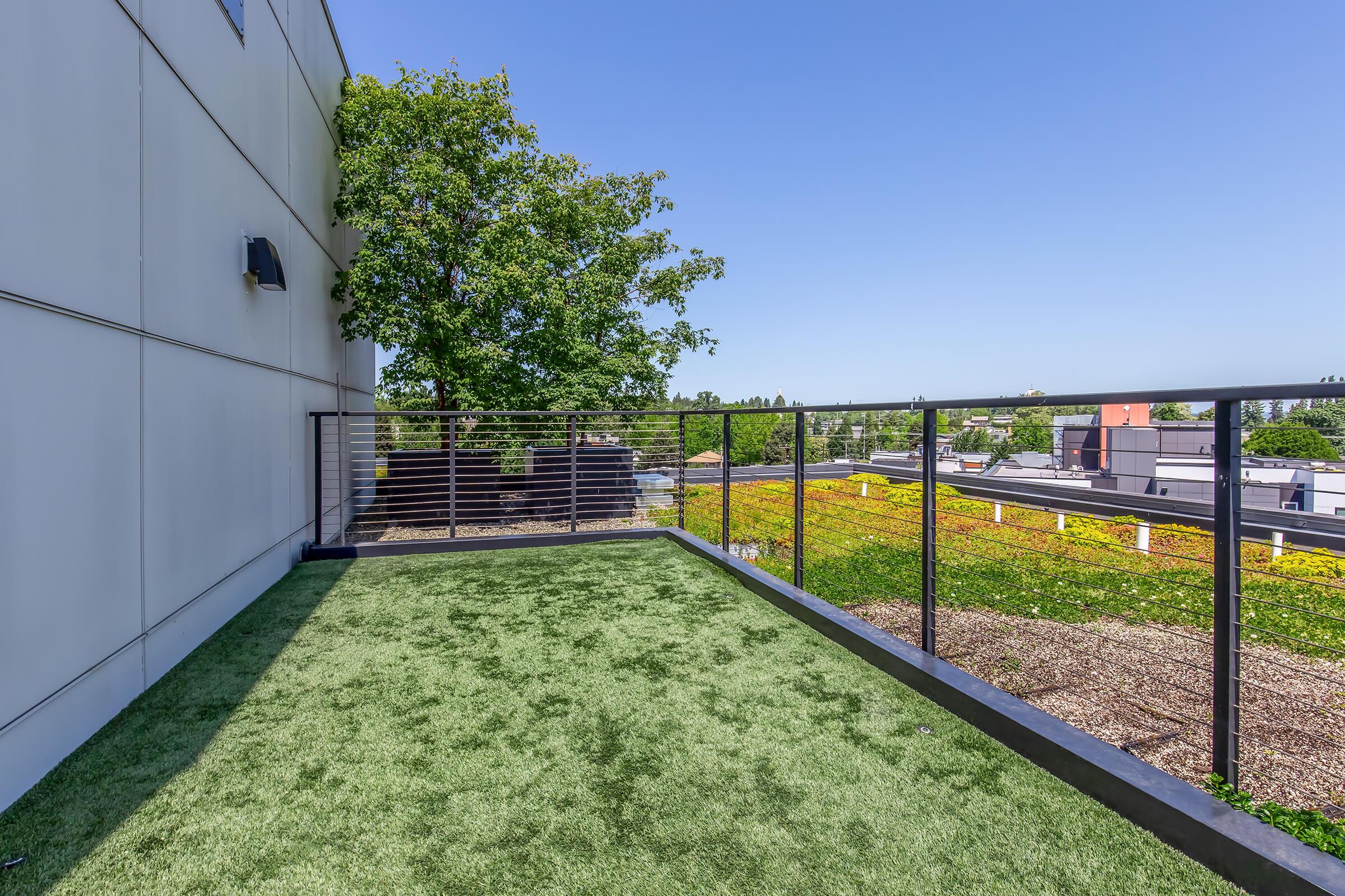 A balcony or terrace with artificial turf, featuring a railing and a tree nearby. The view includes rooftops and green fields under a clear blue sky, indicating a sunny day.