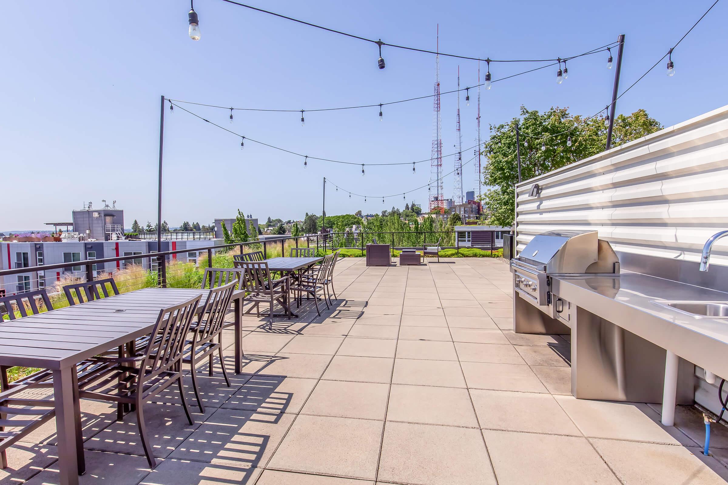 A rooftop terrace featuring outdoor dining tables with wooden chairs, string lights overhead, and a stainless steel barbecue grill. Lush green plants and a partial view of city buildings and communication towers in the background under a clear blue sky.