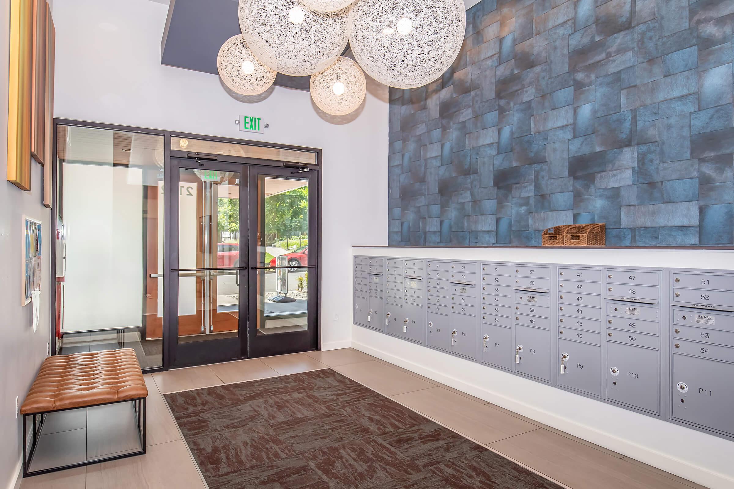 Interior view of a modern lobby featuring a row of metal mailboxes, a decorative bench, and a stylish wall with a textured blue design. Large round light fixtures hang from the ceiling, and there is a glass entrance door leading outside. The space has a clean, contemporary look with neutral colors.