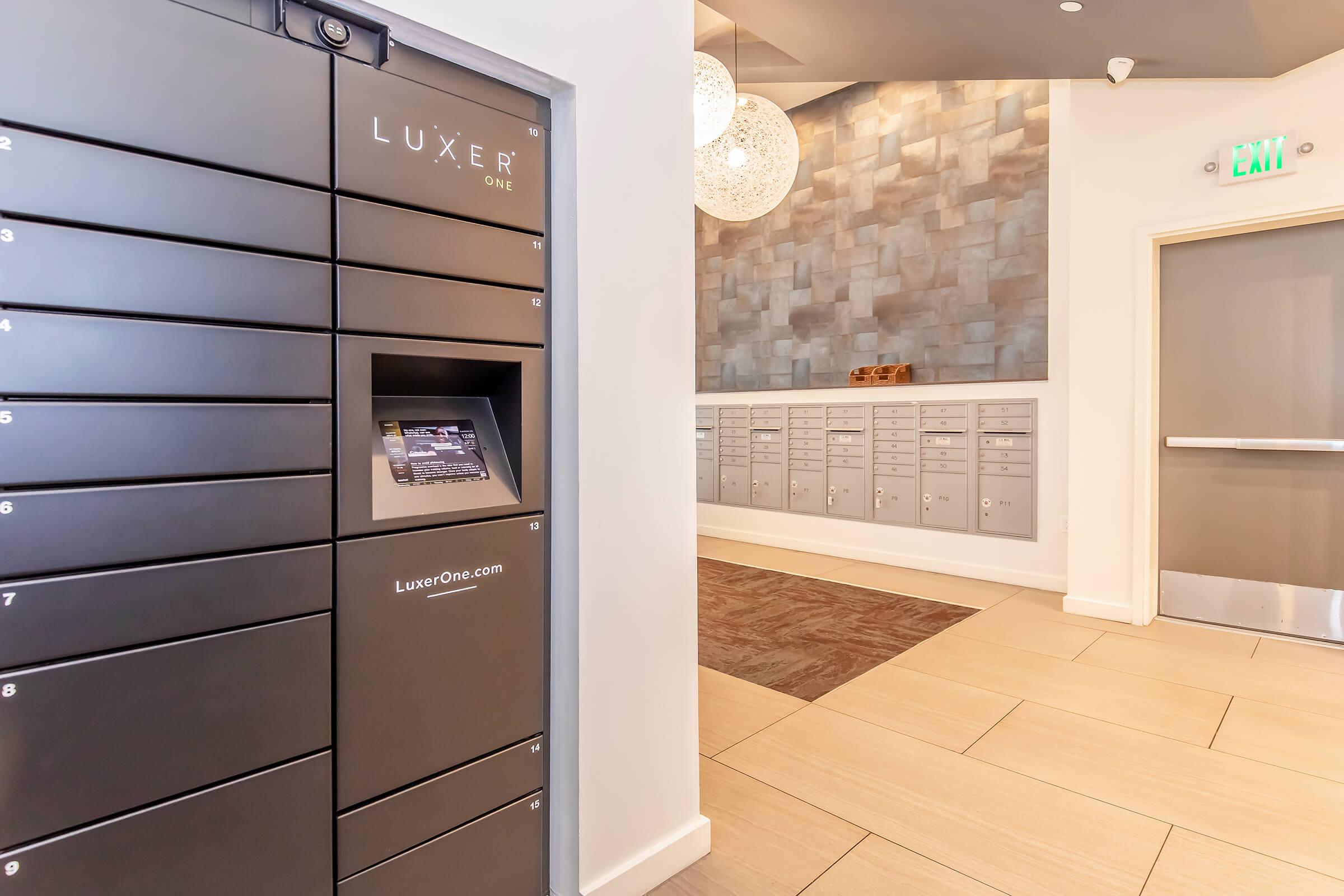 A modern package delivery area featuring a black Luxer One locker system on the left and gray mailboxes on the right. The backdrop includes a textured wall and decorative lighting, creating a sleek and contemporary atmosphere. The flooring is light-colored tiles.