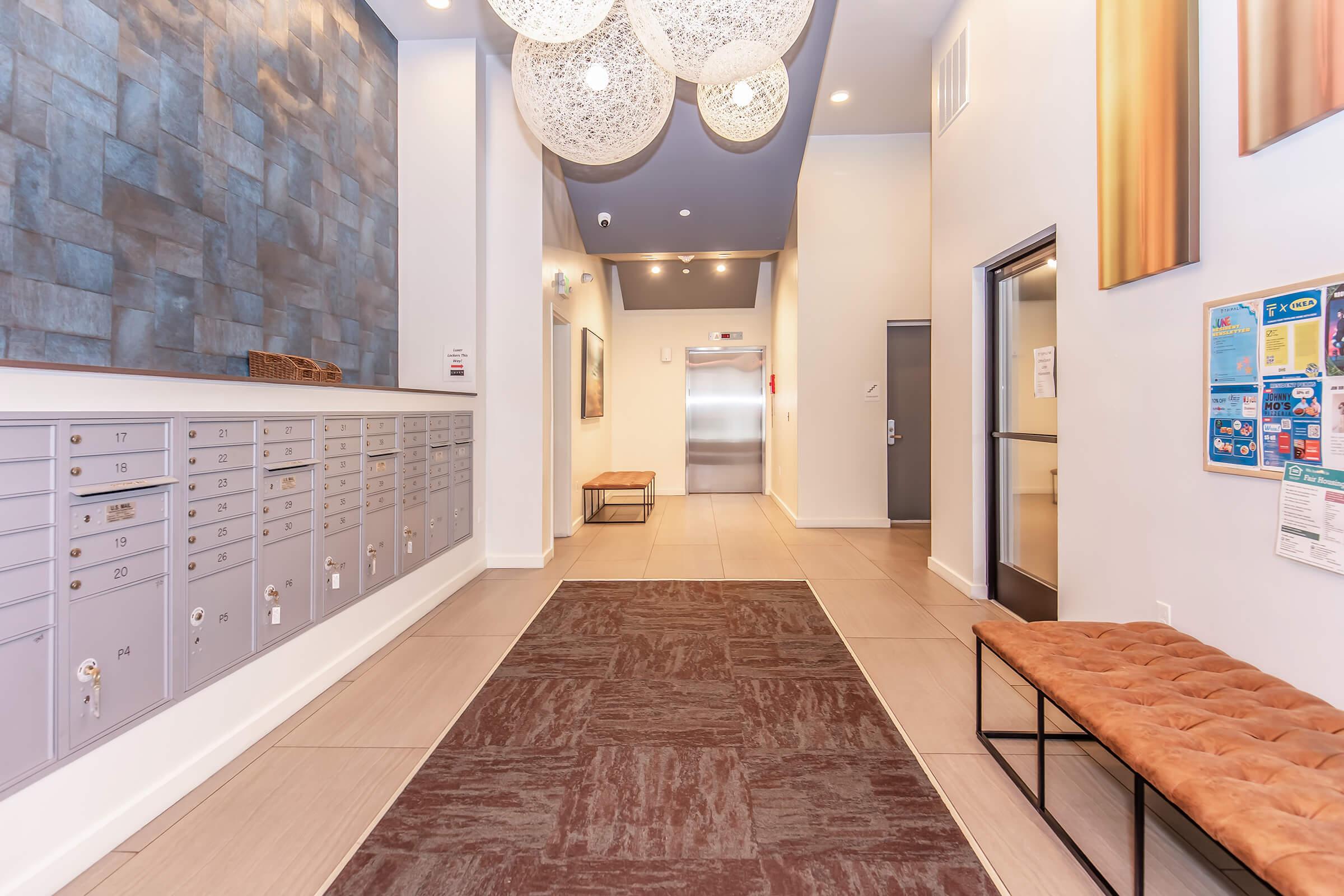 A modern hallway featuring decorative wall panels, a series of pendant lights, and a row of mailbox compartments on the left. There are two benches along the wall and an elevator door at the end of the corridor, with artwork displayed on the walls and a carpet runner leading into the space.