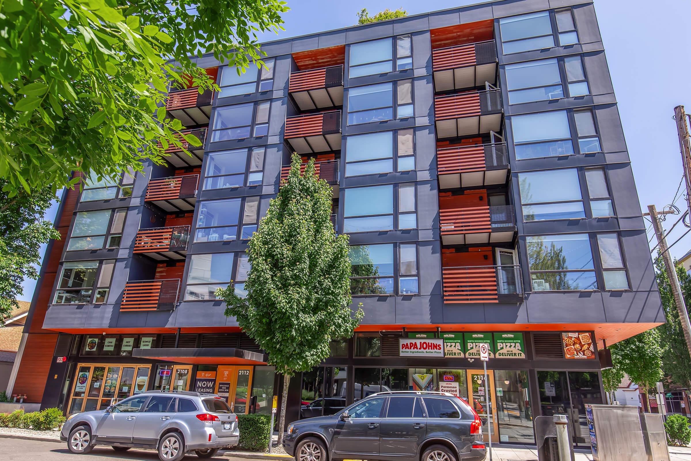Modern multi-story apartment building with large windows and wooden balcony accents. The ground floor features storefronts, including a pizza place, with cars parked in front. Lush green trees are in the foreground, adding to the urban landscape. Clear blue sky overhead enhances the vibrant scene.