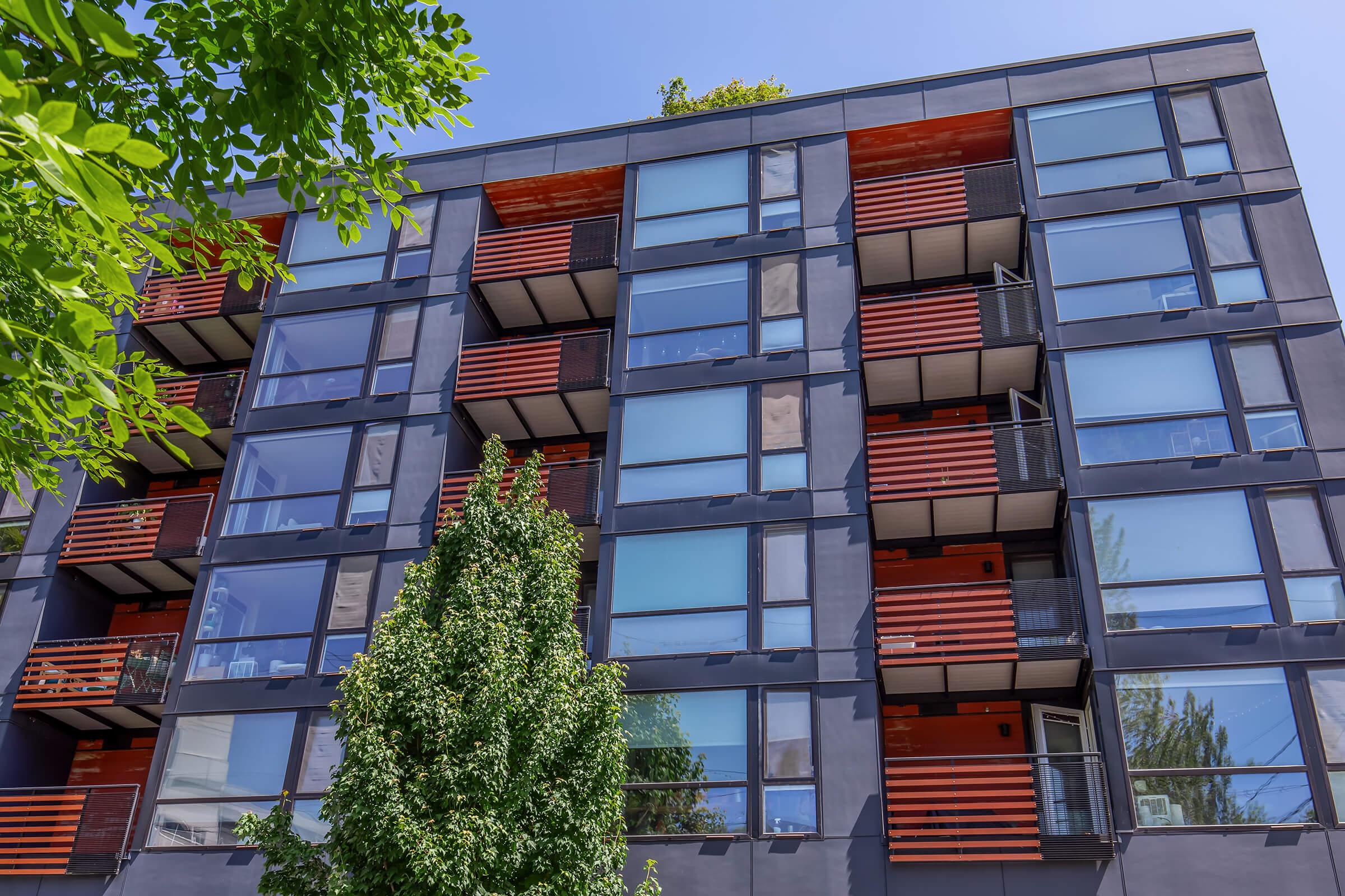 A modern apartment building featuring a sleek facade with large windows and red wooden balcony accents. Lush green trees frame the view, contrasting with the contemporary design of the structure, which has multiple stories and a vibrant, urban feel.
