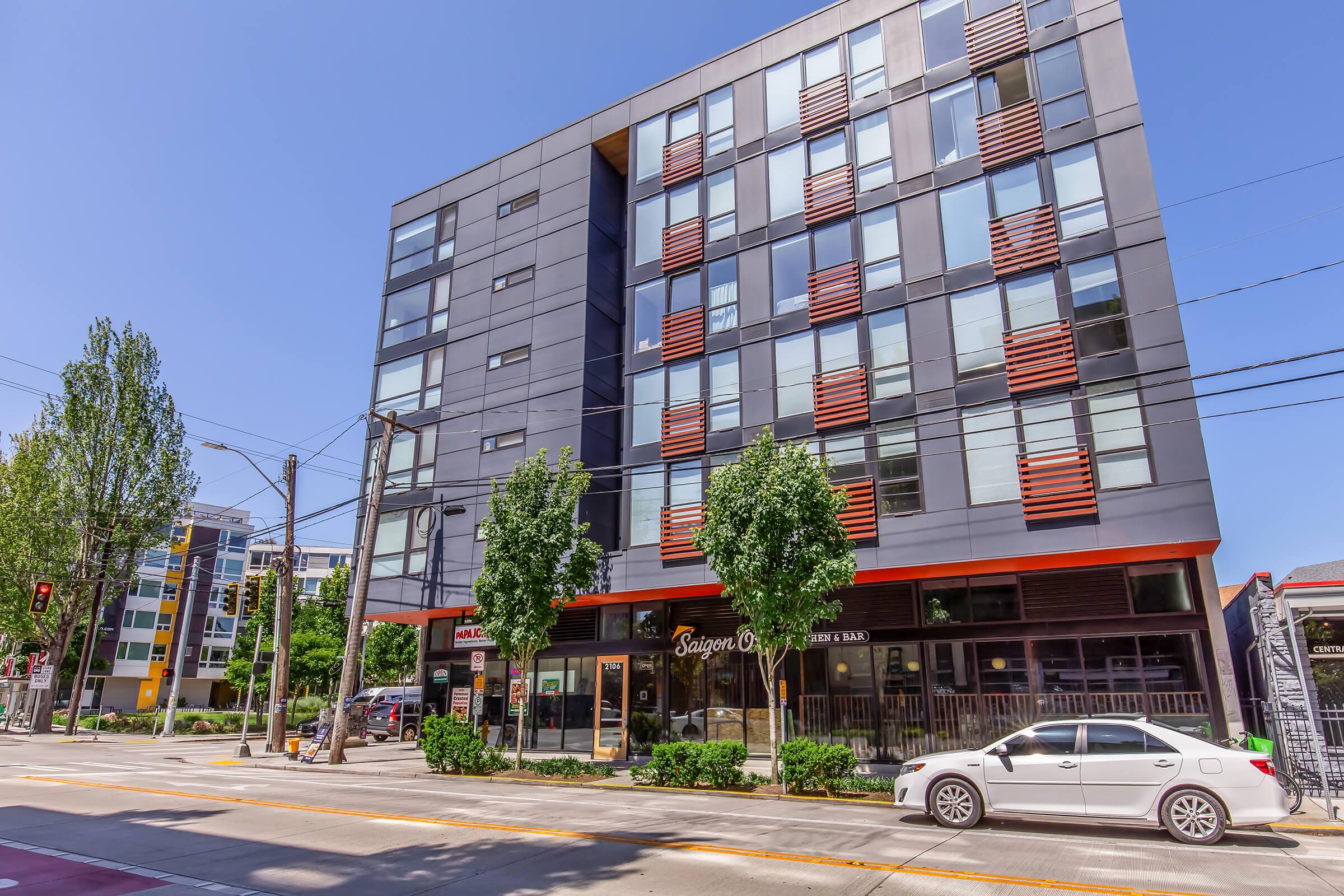 A modern multi-story building featuring large windows and a mix of dark and light facade elements. The ground floor has commercial space with signage for a restaurant or bar. Surrounding the building are trees, and a white car is parked beside the street in a busy urban setting.
