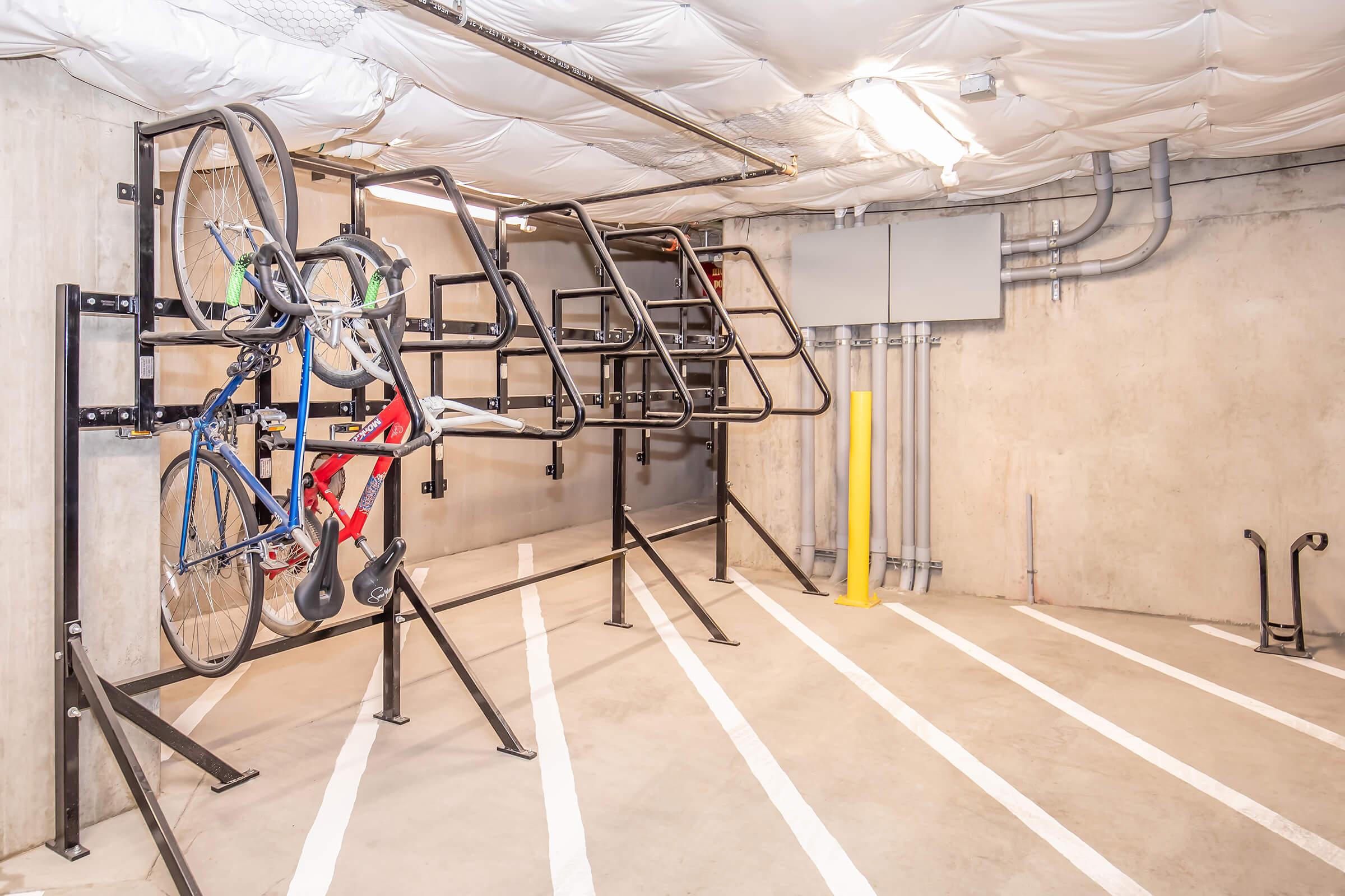 A modern bike storage area featuring vertical racks for bicycles. Two bikes are mounted on the racks, one blue and white, and the other red and white. The space has a concrete floor and walls with visible electrical conduits and a yellow post. The area is well-lit and organized, with clear lines marking storage spots.