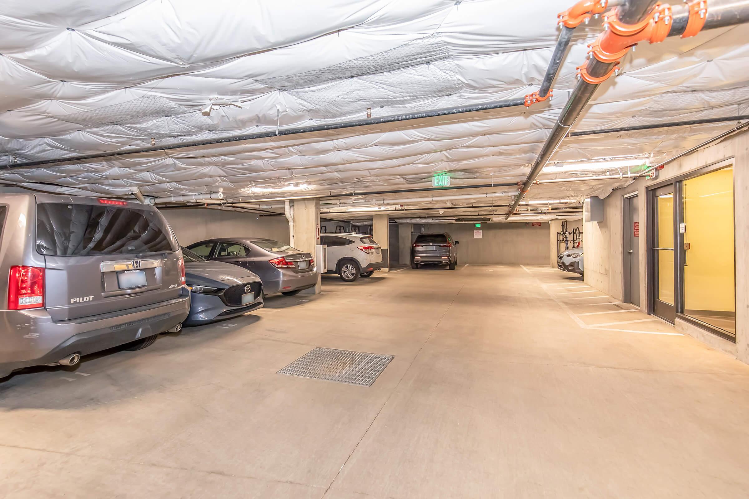 A well-lit underground parking garage with multiple parked vehicles, including a gray SUV and a silver sedan. The ceiling has exposed pipes and a smooth concrete floor. There are designated parking spaces, along with an exit door visible at the far end. Soft lighting enhances the overall visibility of the space.