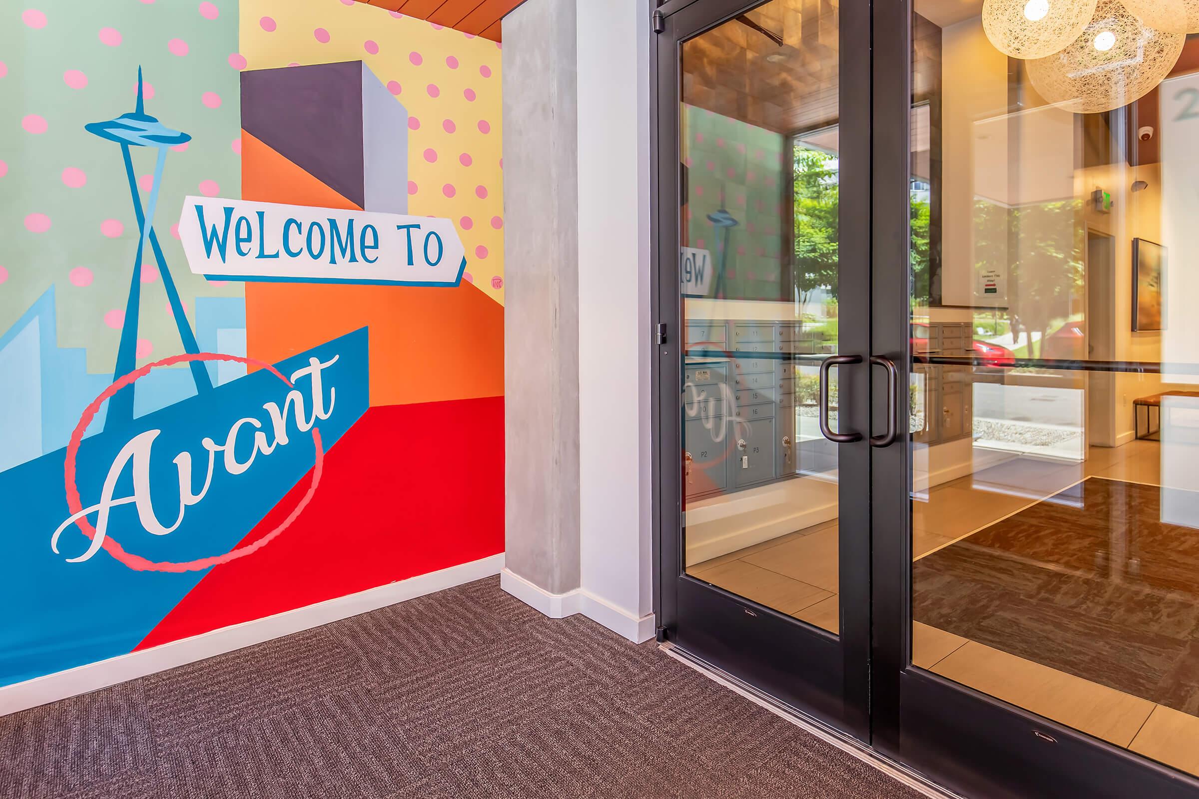 Colorful entrance area featuring a mural that says "Welcome to Avant," with geometric designs and a silhouette of the Space Needle in Seattle. The wall is painted in vibrant colors like red, blue, and orange, contrasting with a modern glass door leading to the interior. The flooring is a neutral carpet.