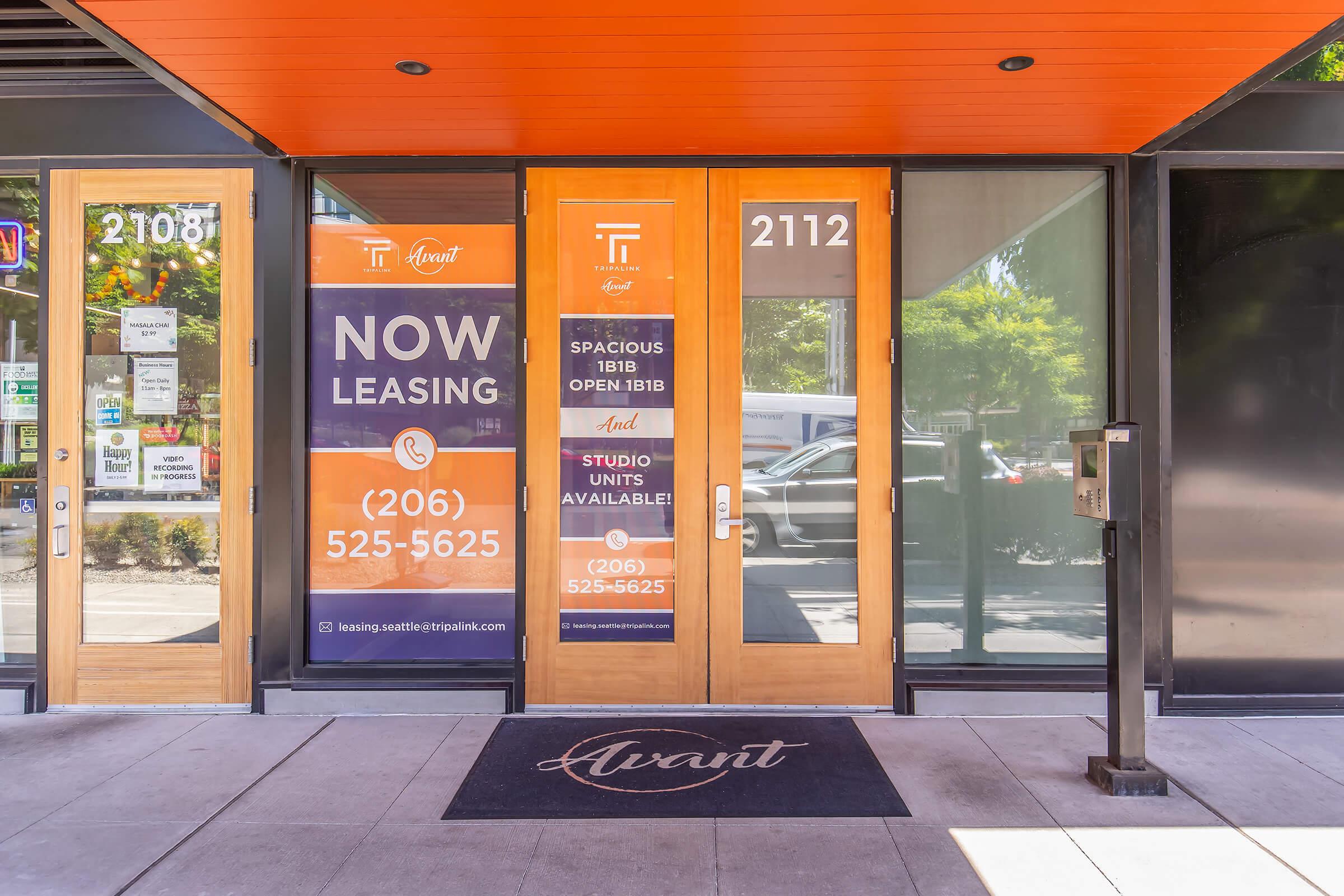 Front entrance of a leasing office with large glass doors displaying signs. The left door has a sign stating "NOW LEASING" with a phone number for inquiries. The right door features information about available studio and one-bedroom units. A welcome mat is in front, and there's a mailbox on the side.