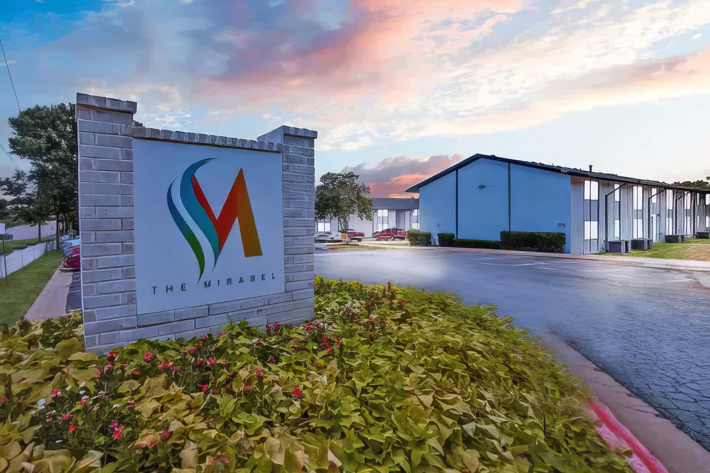 Sign for "The Mirabell" with a colorful logo, surrounded by landscaped flower beds. In the background, a multi-unit building can be seen against a pastel sky at dusk. The setting appears well-maintained, conveying a welcoming atmosphere.