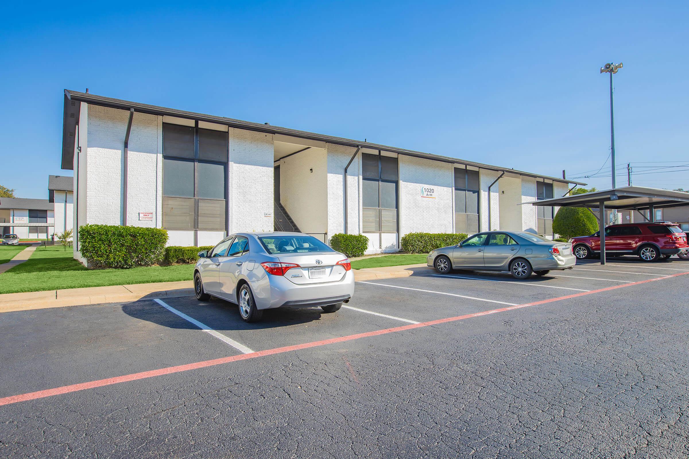 A parking lot in front of a multi-story residential building. The building has a white exterior with large windows and is surrounded by well-maintained grass. Several cars are parked in the lot, including a silver sedan, with clear blue skies overhead.