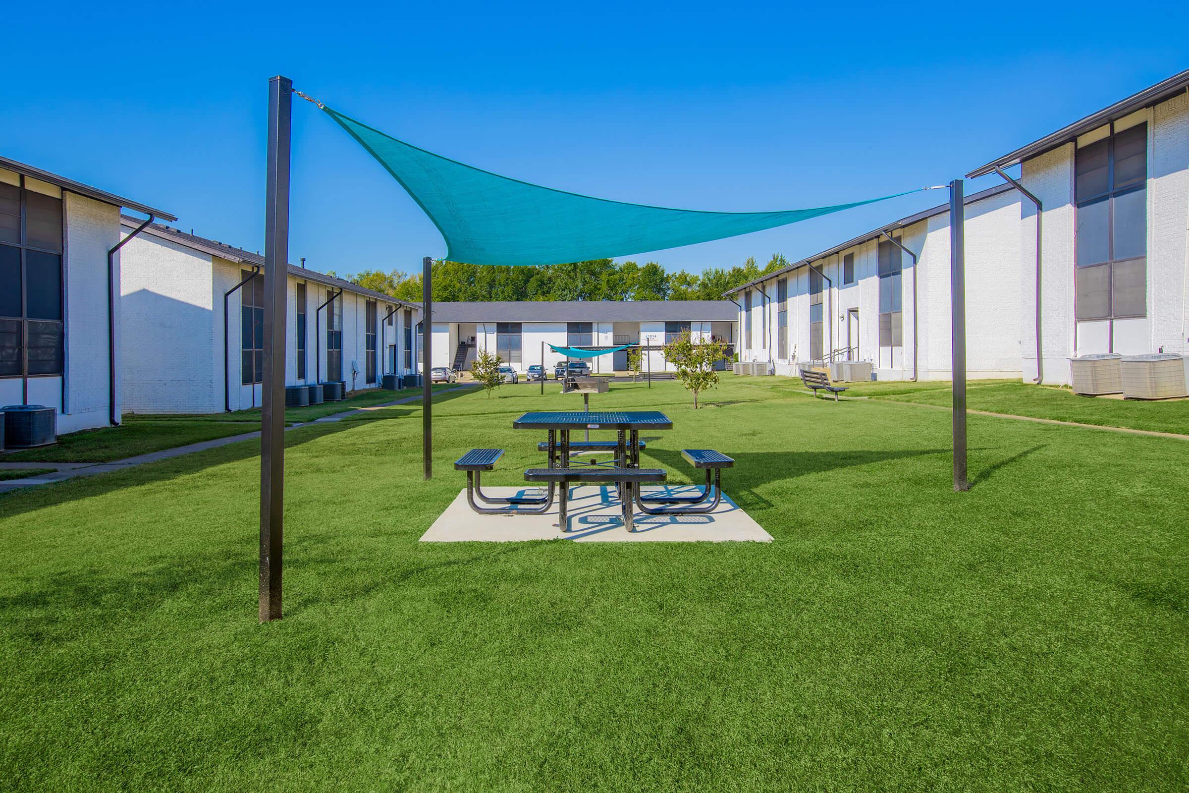 A bright outdoor area featuring a green lawn with a shaded picnic table beneath a blue canopy. Surrounding the area are two buildings with white exteriors, and clear blue skies above, creating a welcoming atmosphere for relaxation and social gatherings.