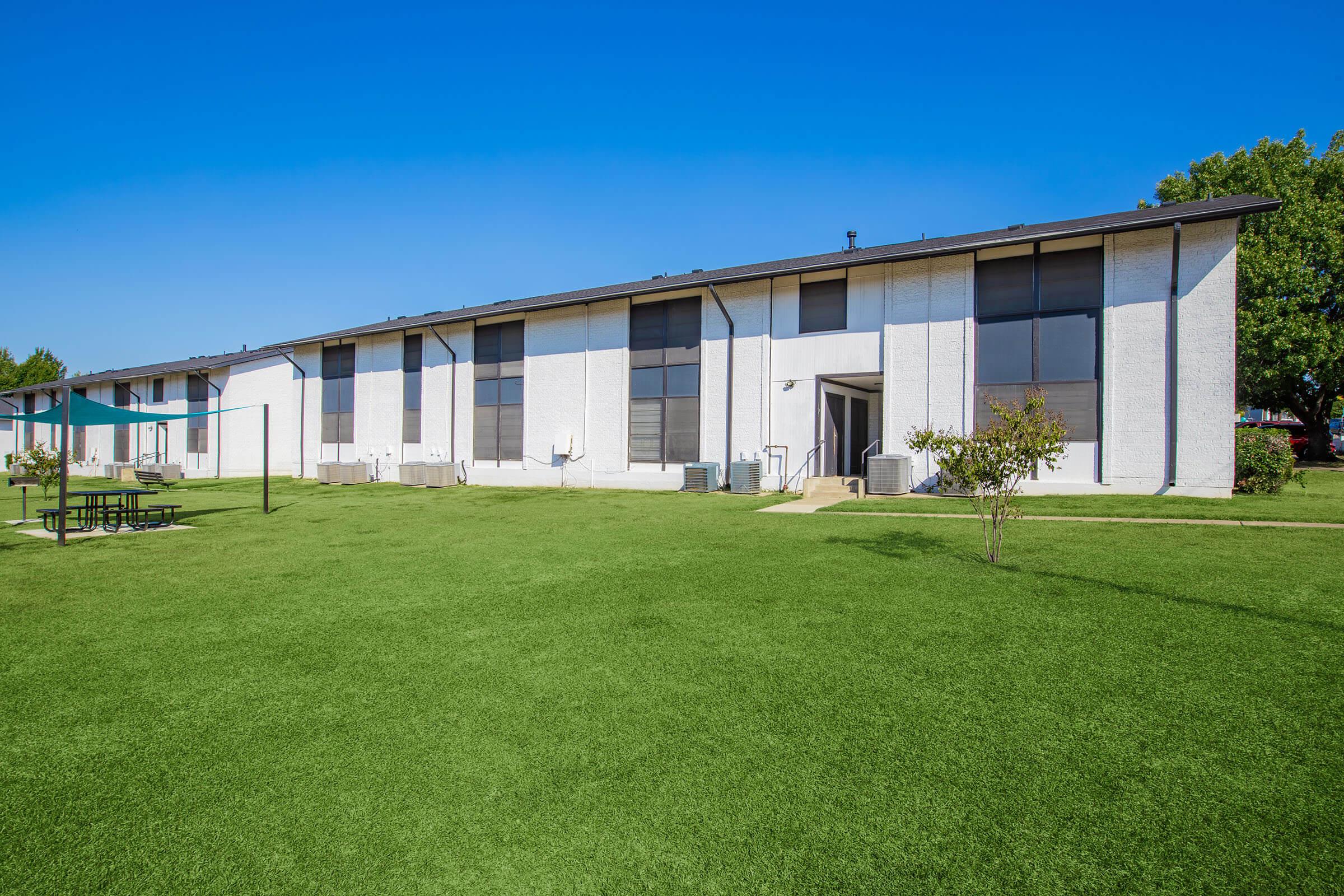 A modern one-story residential building with large windows, set on a grassy area. In the foreground, there are picnic tables and a shaded area with a sail. The sky is clear and blue, creating a bright and inviting atmosphere.