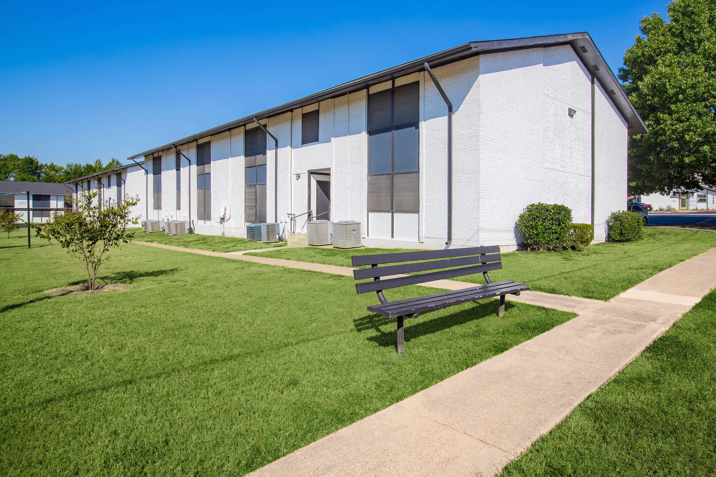 A wide shot of a residential building with a white exterior, large windows, and air conditioning units, surrounded by well-maintained green grass. A black bench is positioned along a path leading to the entrance, under a clear blue sky. A tree stands nearby, adding to the outdoor ambiance.