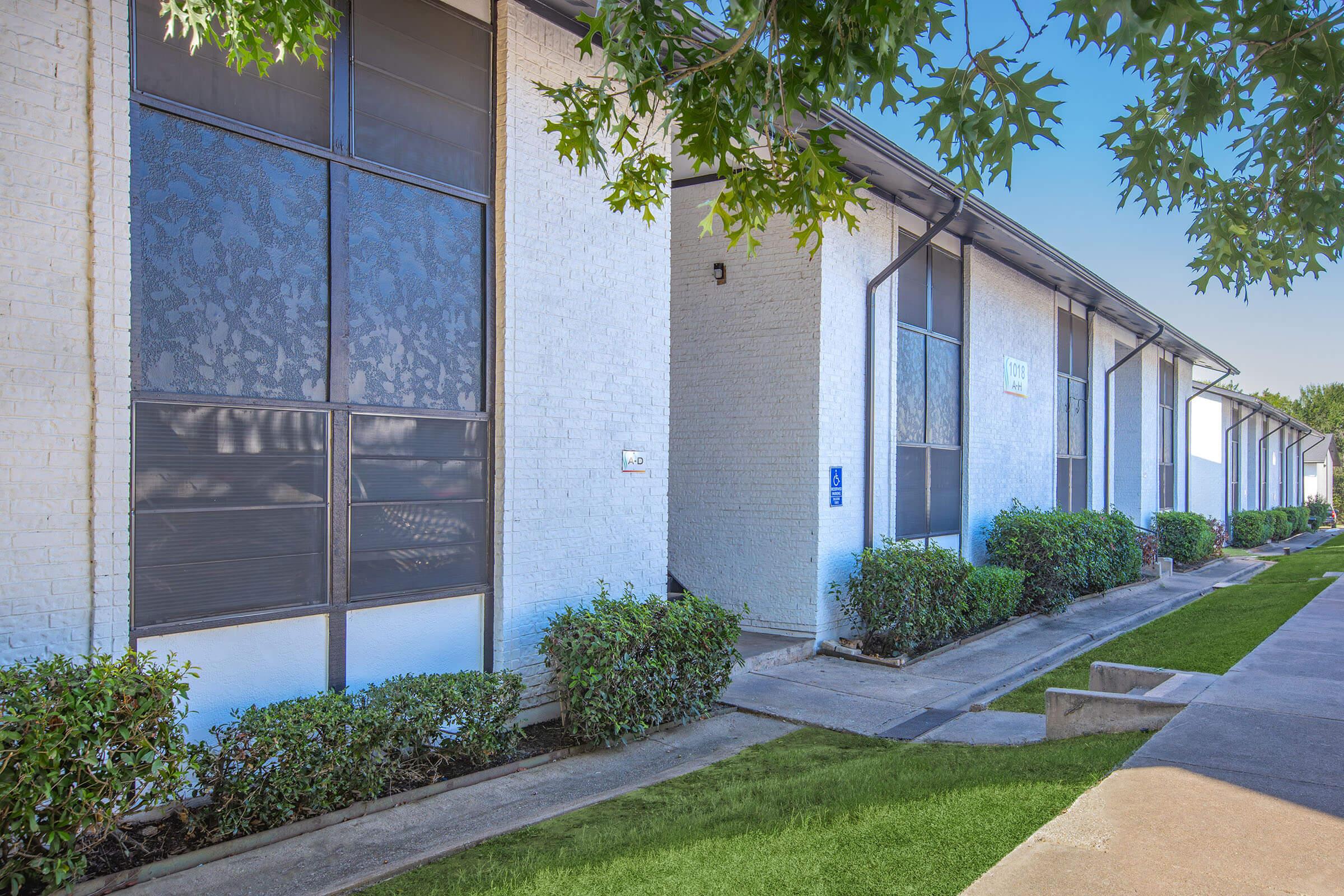 A row of low-rise apartment buildings with white exteriors and large windows. The walkway is lined with neatly trimmed shrubs and grass, leading to each entrance. The scene is bright and well-maintained, with trees providing shade in the foreground.