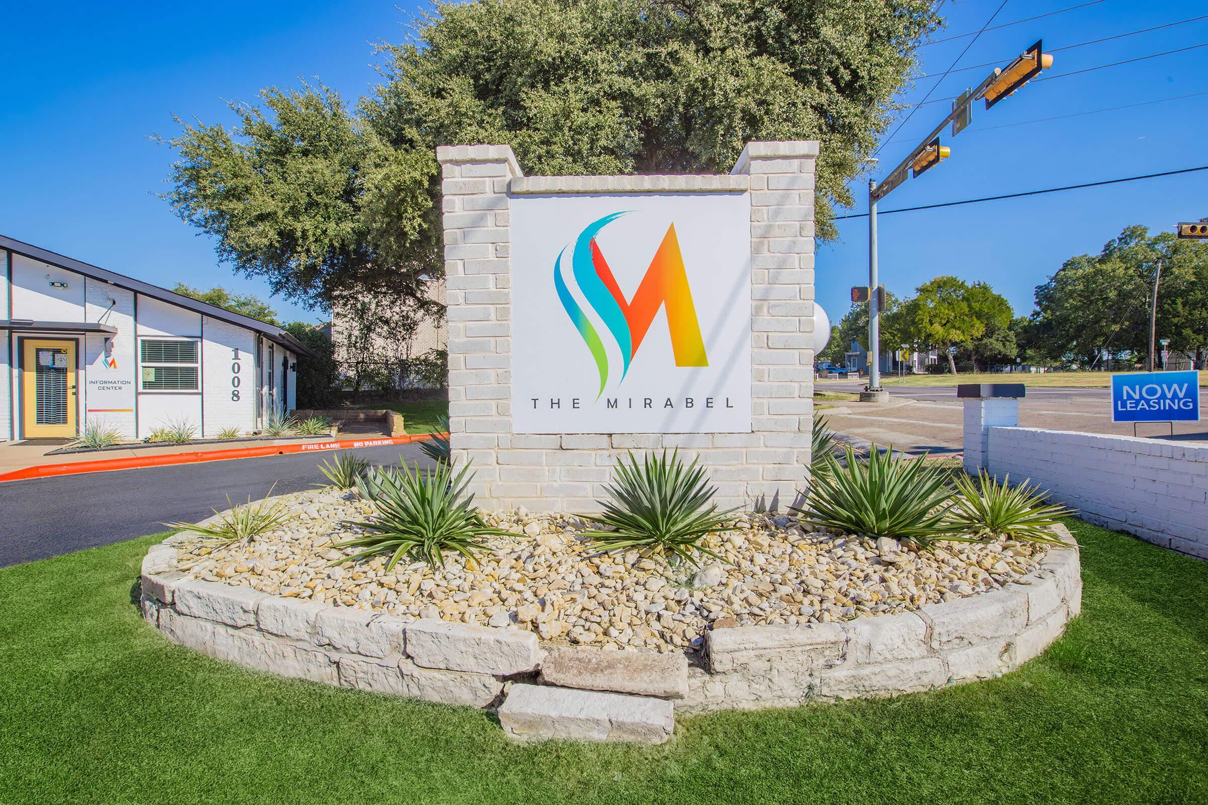 Sign for "The Mirabel" featuring a colorful logo on a stone pedestal surrounded by decorative plants and gravel. The background includes a paved area and traffic lights. A "Now Leasing" sign is visible nearby.