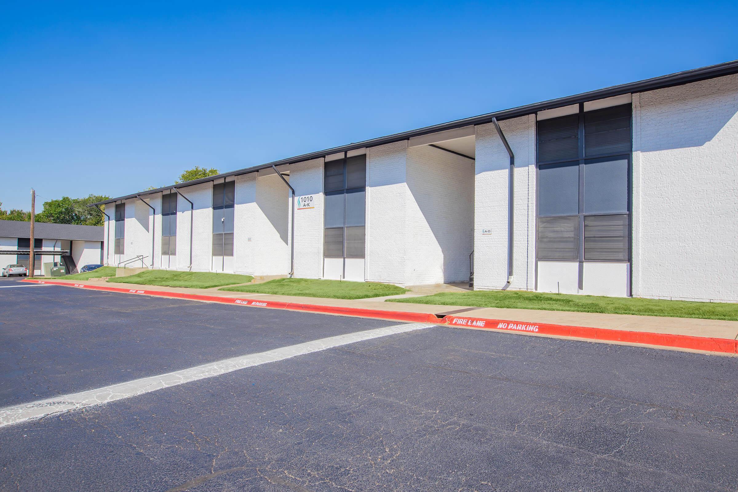 Exterior view of a two-story white apartment building with large windows and a flat roof. The building is set back from the road, with a red curb indicating no parking. A landscaped area with green grass is visible in front of the building under a clear blue sky.