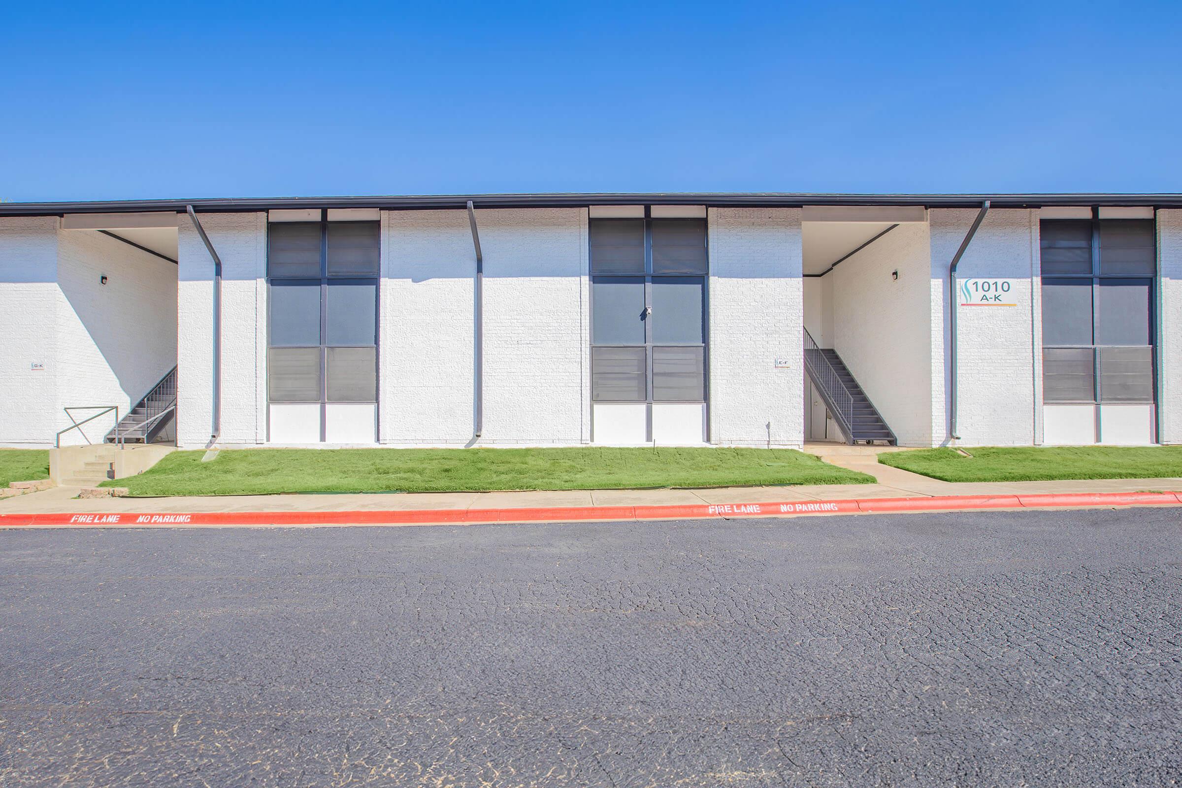 A two-story residential building with a white exterior and large windows. The entrance features a staircase leading to the second floor. Green grass lines the front, while a paved road is visible in front of the building. The sky is clear and blue.