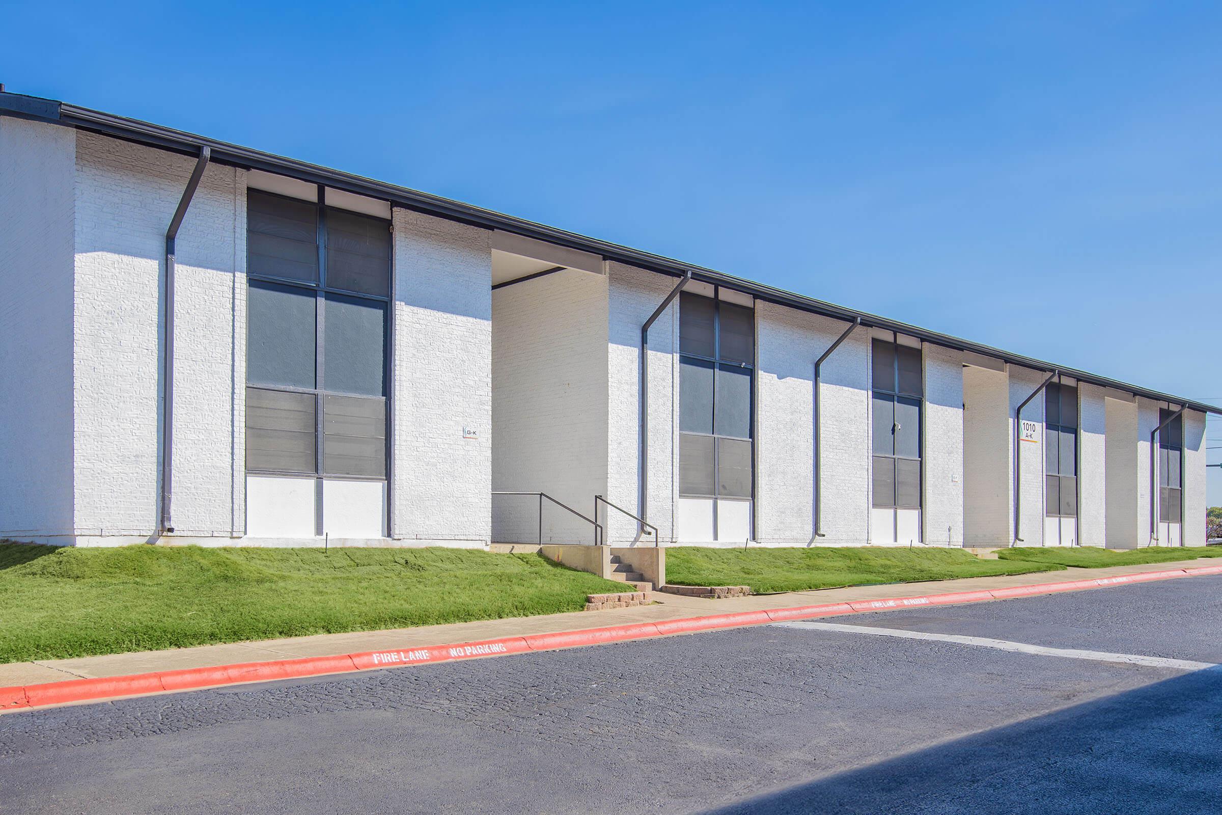 Exterior view of a modern, white apartment building featuring large windows and a pathway leading to the entrance. The building is set against a clear blue sky, with green grass in front and a red curb along the driveway.