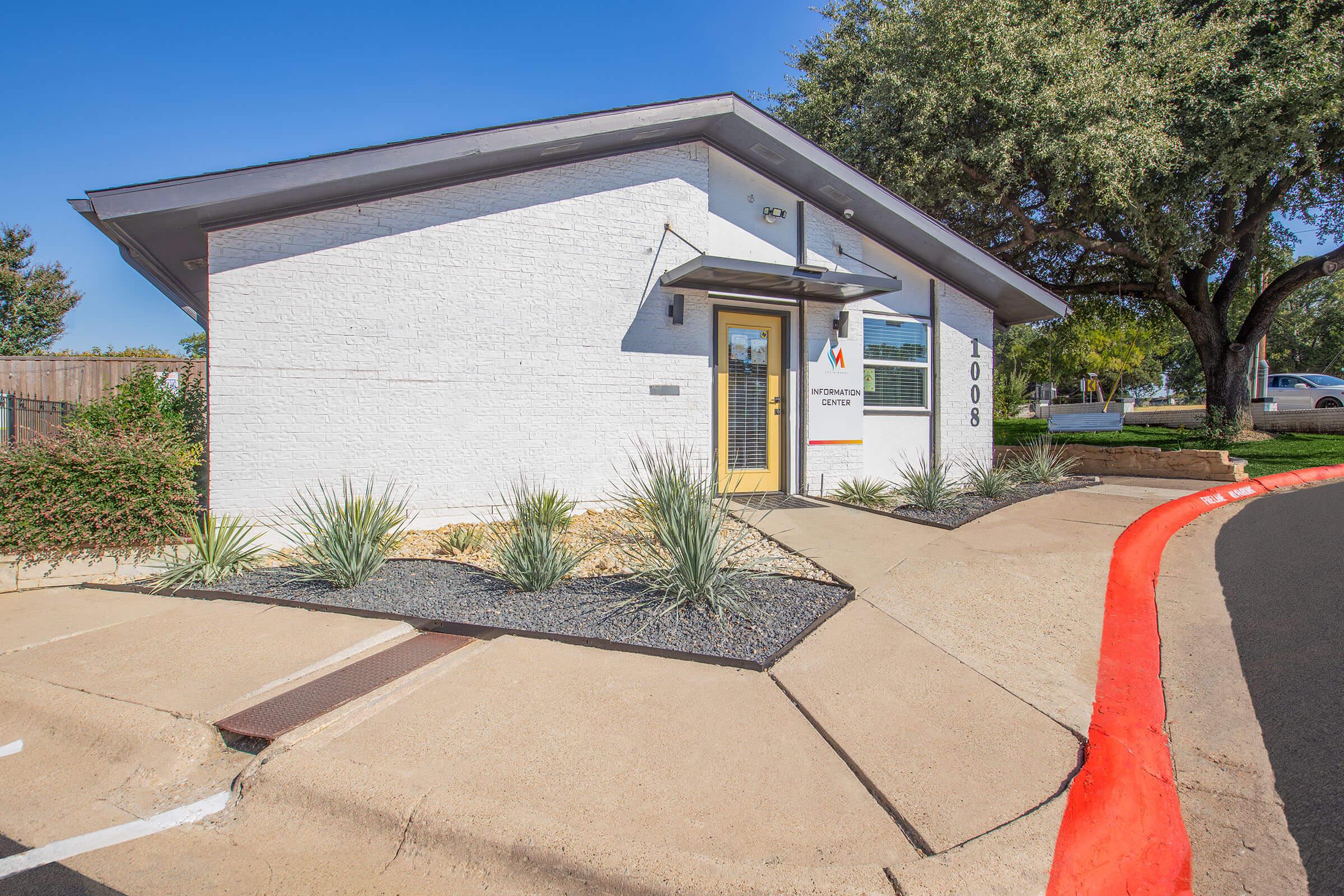 A modern white building with a flat roof, featuring large windows and a welcoming entrance. The exterior is surrounded by low-maintenance landscaping, including small shrubs and decorative stones. A red-painted curb leads up to the entrance, indicating parking or access. Clear blue sky above.