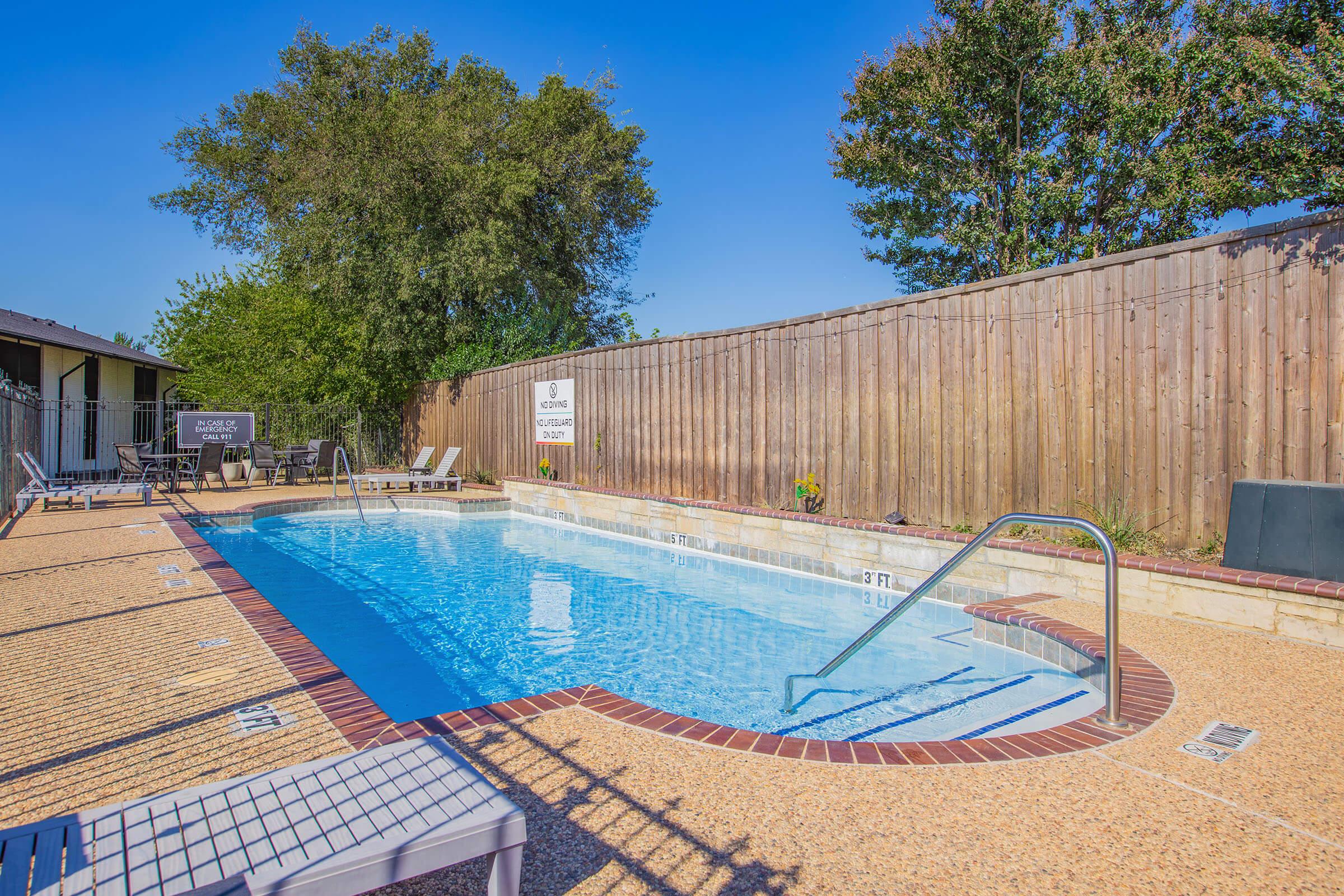 A serene, inviting swimming pool surrounded by a wooden fence. The pool features a ladder for easy access and is complemented by lounge chairs arranged nearby. A clear blue sky provides a bright backdrop, enhancing the relaxing atmosphere of this outdoor space.