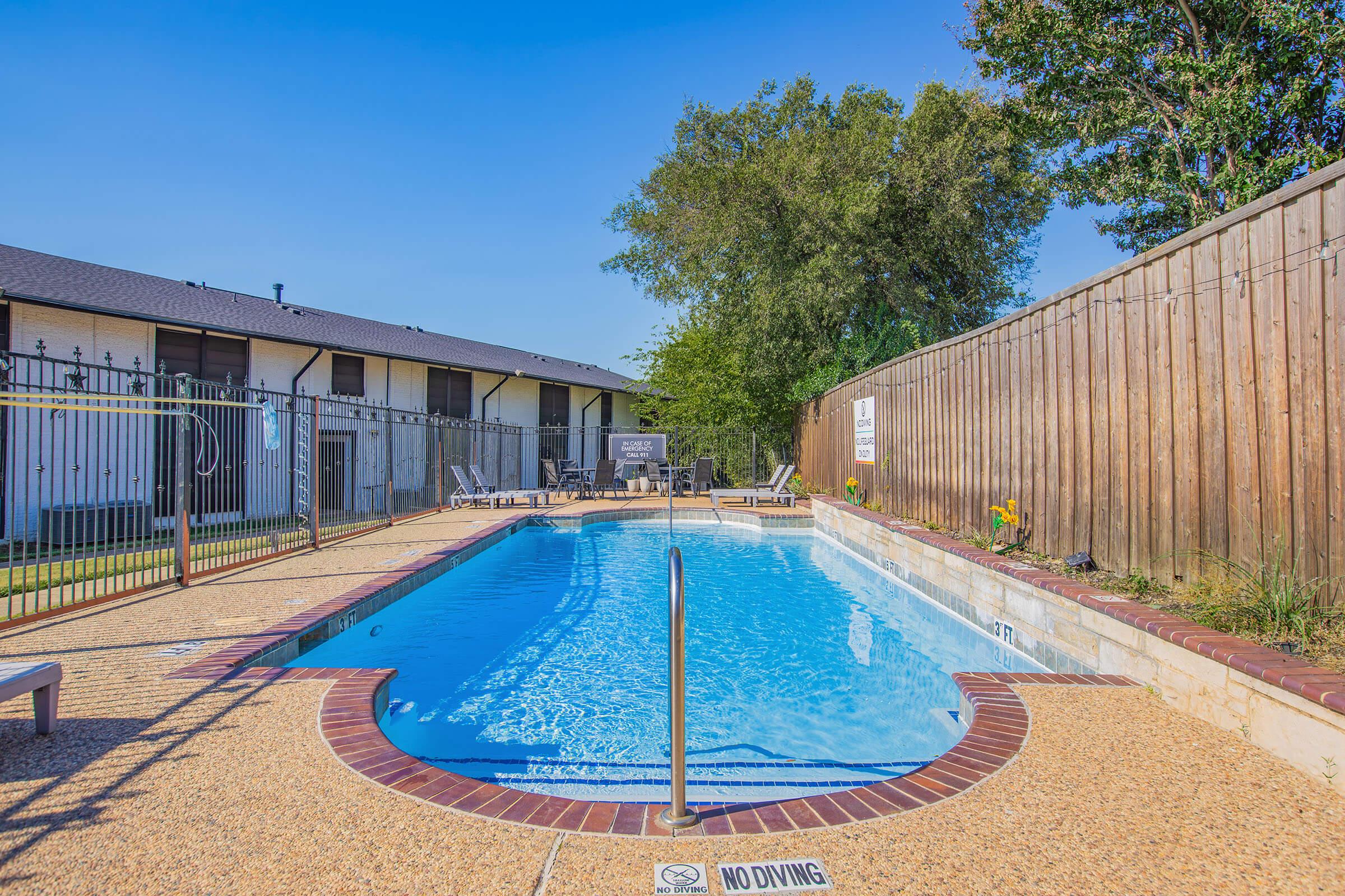 A clear swimming pool with a ladder, surrounded by a fenced area. The pool area features lounge chairs and potted plants. A wooden fence provides privacy, and there is a "No Diving" sign near the pool's edge. Bright blue sky and greenery in the background complete the scene.