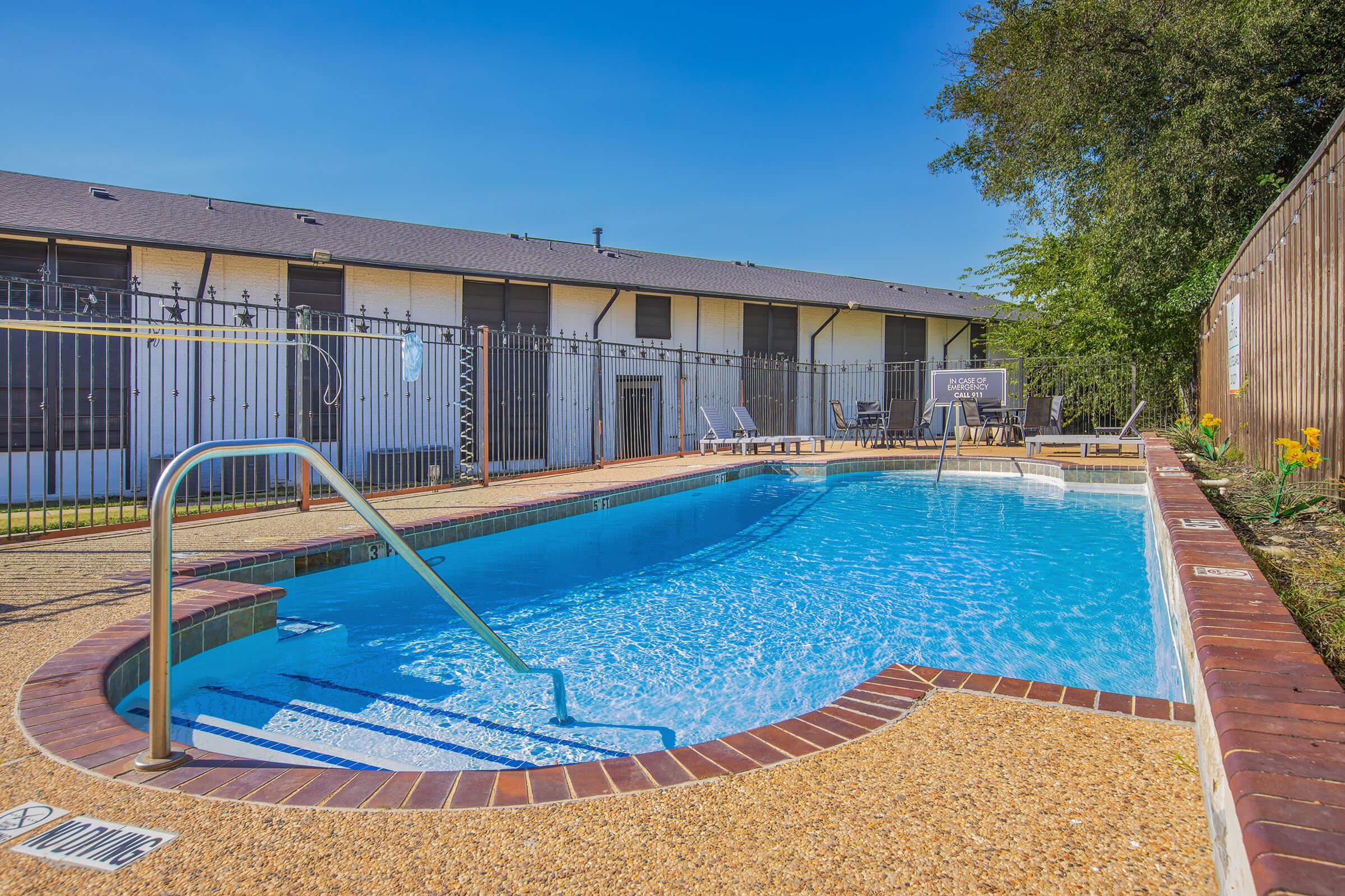 A clear swimming pool surrounded by a fenced area, with lounge chairs positioned nearby. The pool features a set of stairs leading into the water, and there are green plants and flowers along the perimeter. In the background, there are apartment buildings under a bright blue sky.
