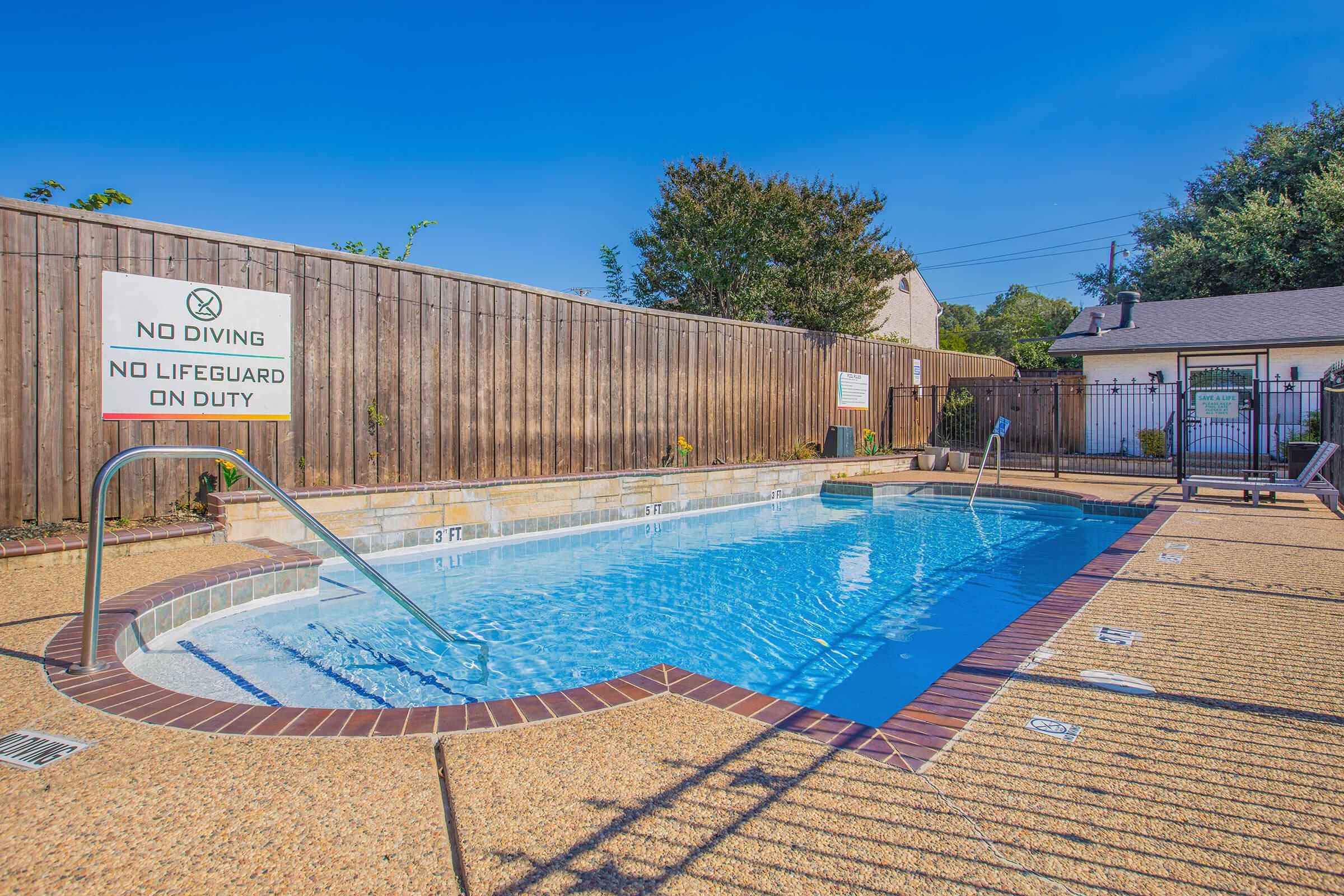 A small outdoor swimming pool with clear blue water and a textured concrete deck. A wooden fence surrounds the pool area, featuring a sign that reads "No Diving" and "No Lifeguard on Duty." The setting is sunny and includes a few green plants in the background.