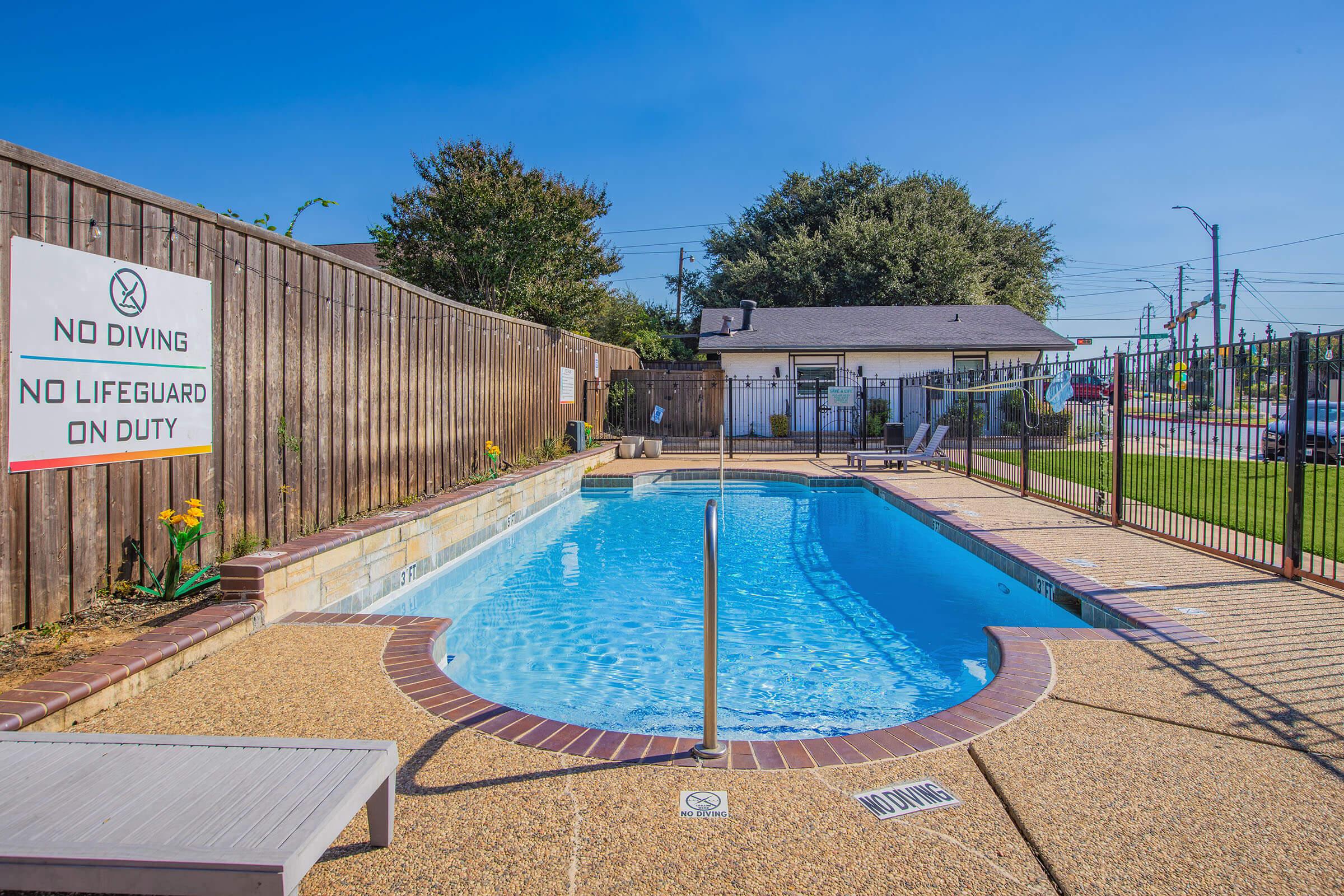 A clear swimming pool surrounded by a fenced area, with a "No Diving" sign posted. There are lounge chairs beside the pool, colorful flowers near the fence, and a residential building in the background under a bright blue sky. The area appears sunny and inviting.