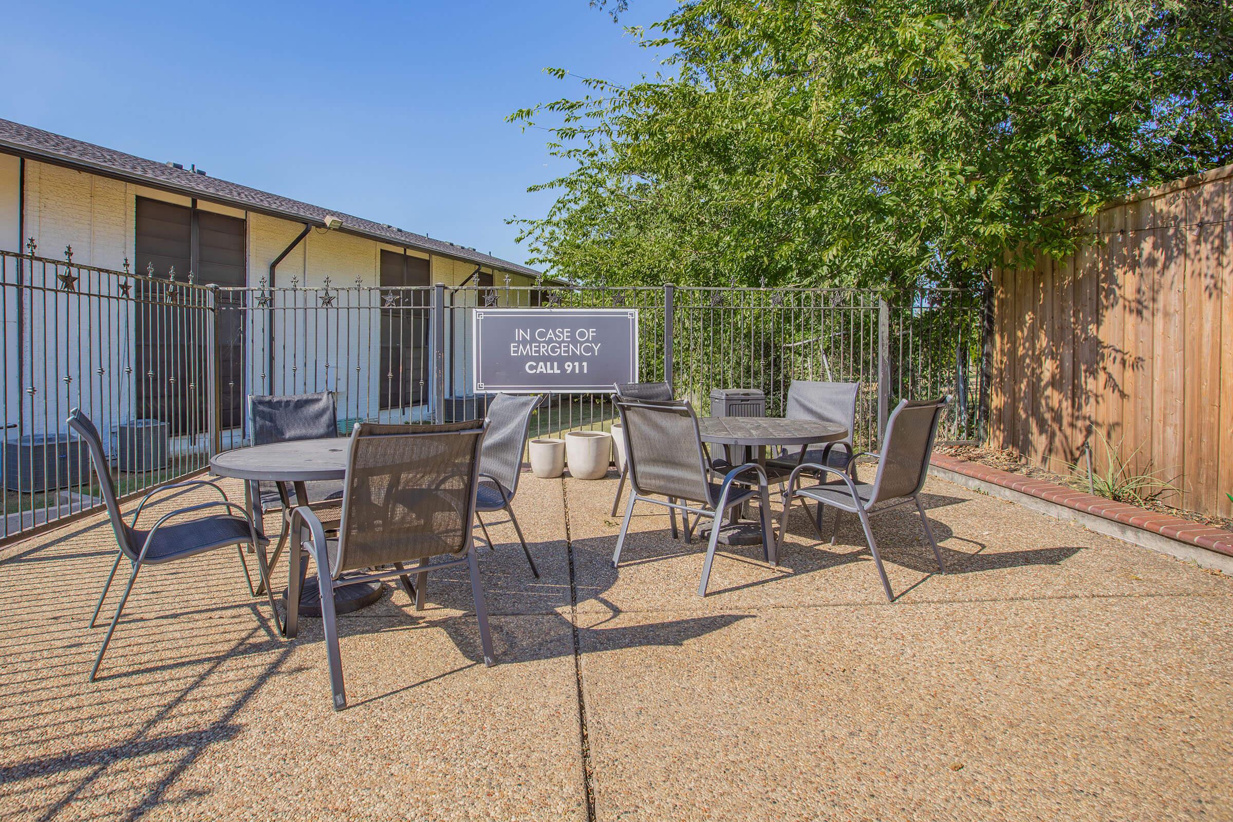 Outdoor patio area with a concrete surface, featuring several metal tables and chairs. A sign stating "In case of emergency call 911" is prominently displayed. The background includes a fence and some greenery, suggesting a well-maintained environment.