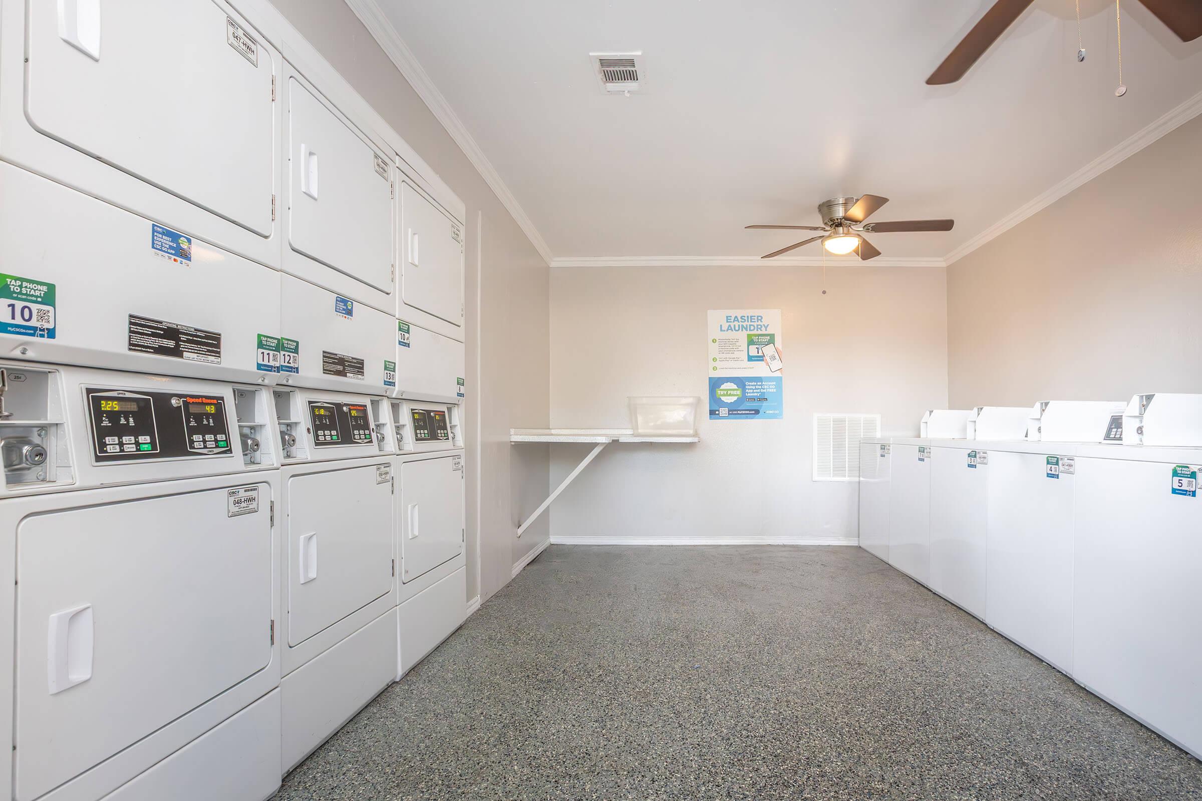A clean and well-lit laundry room featuring several stacked washing machines and dryers along one wall. There is a folding table and a ceiling fan, with informational posters on the wall. The floor is gray and polished, creating a neat and organized atmosphere.